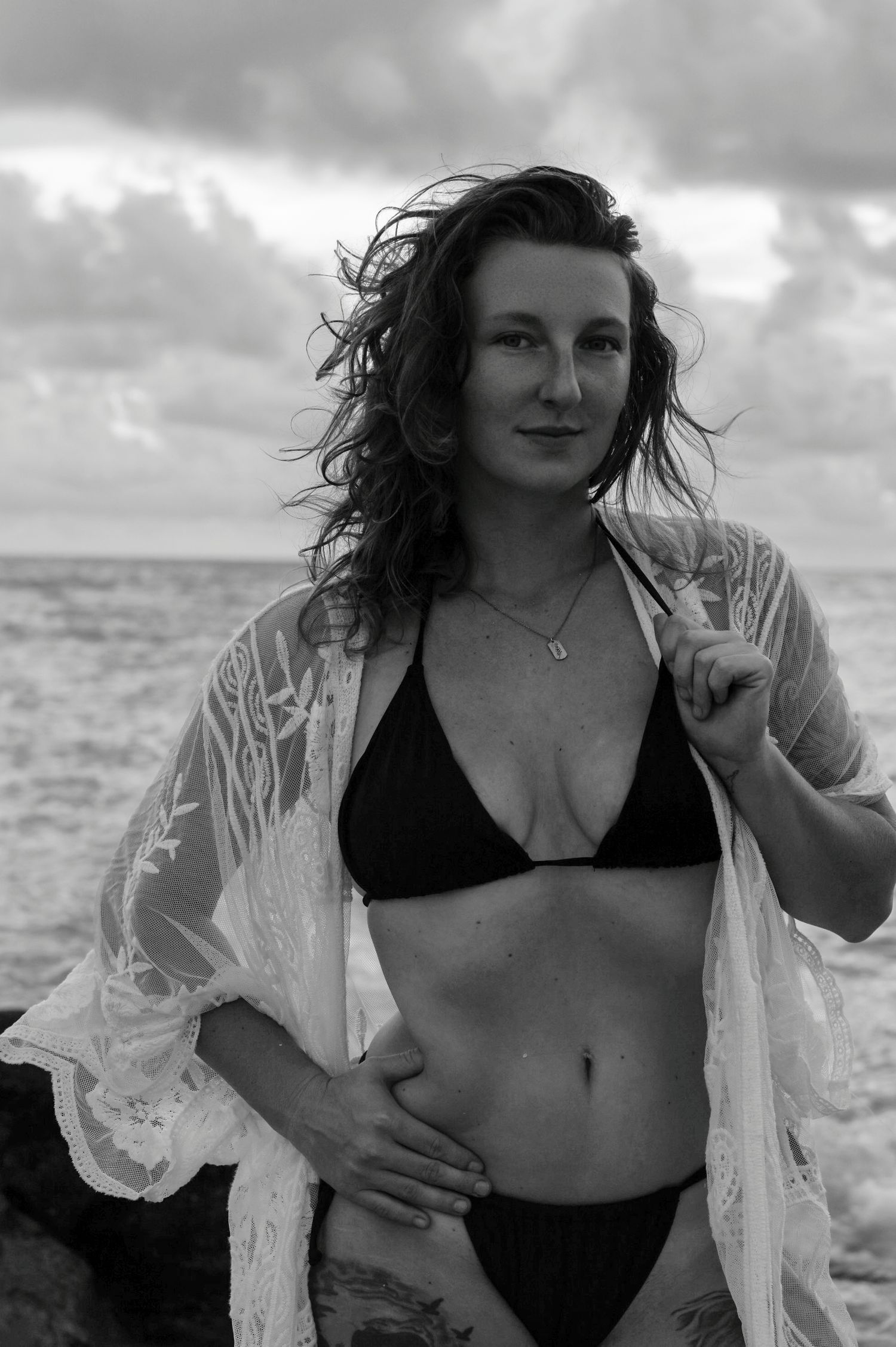 Black and white beach photo of a woman in bikini and lace cover-up with wind-blown hair by the ocean.