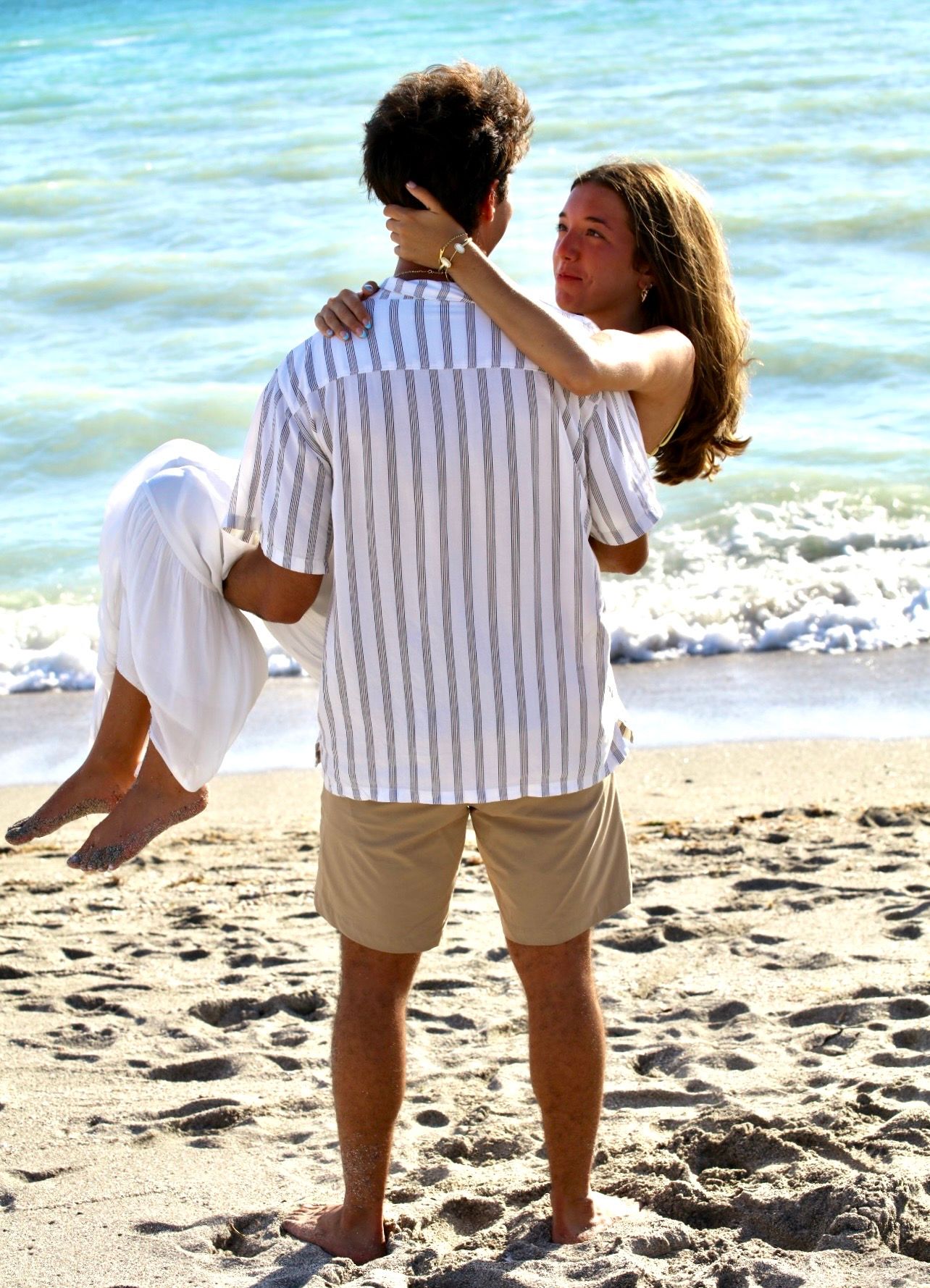 A man carries a woman in his arms on a sandy beach, as she wraps her arms around his neck near the ocean.