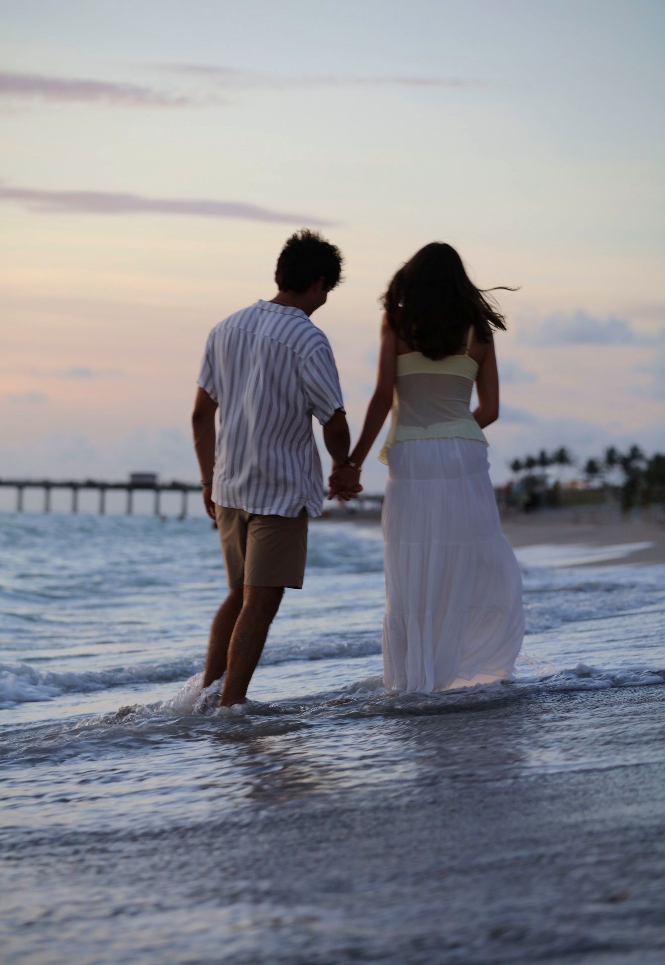 A couple walks hand in hand along the shoreline at sunset near a beach pier.