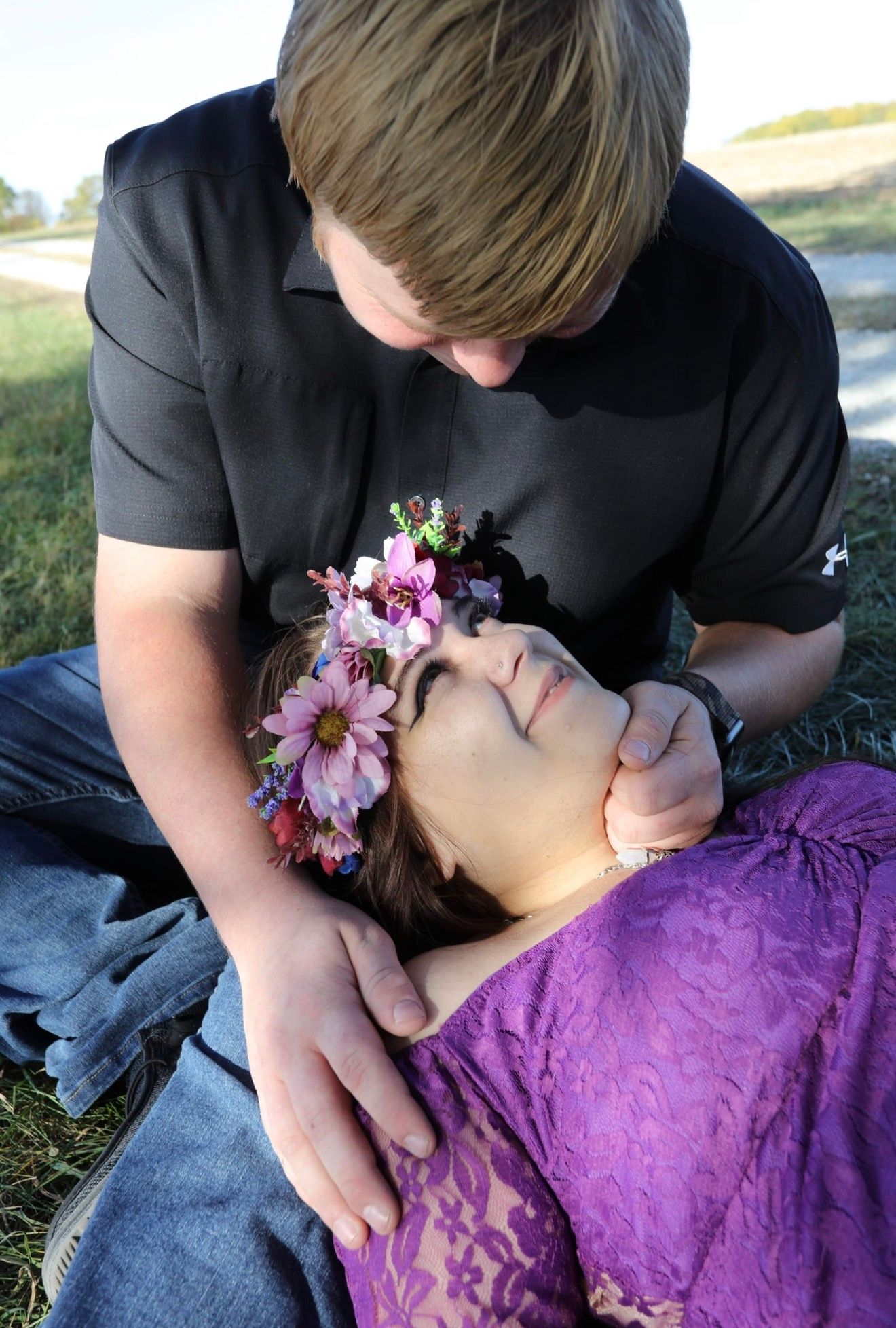 A young couple shares a tender moment outdoors, woman in purple lace dress with floral crown lying in mans arms.