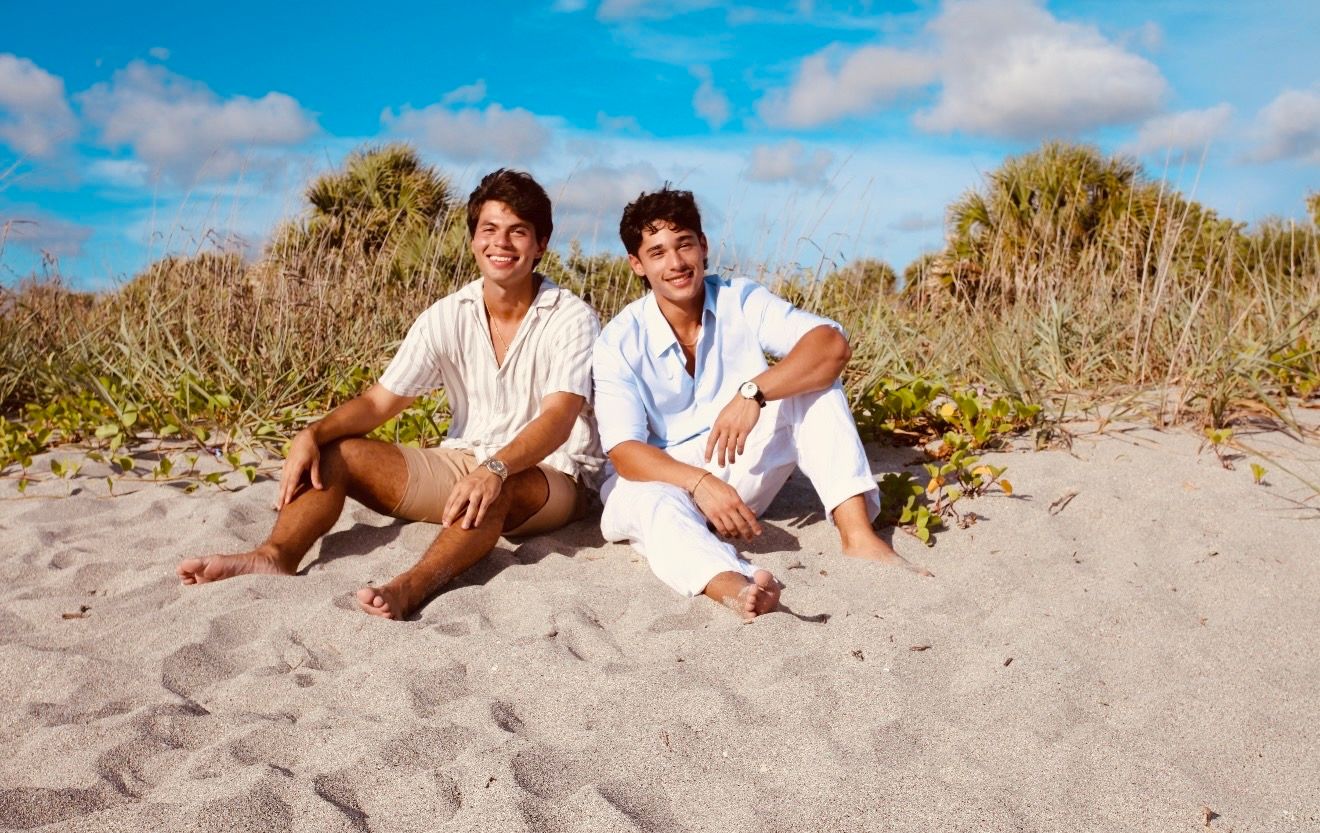 Two young men sitting together on sandy beach, wearing white casual outfits, smiling with tropical greenery behind them.
