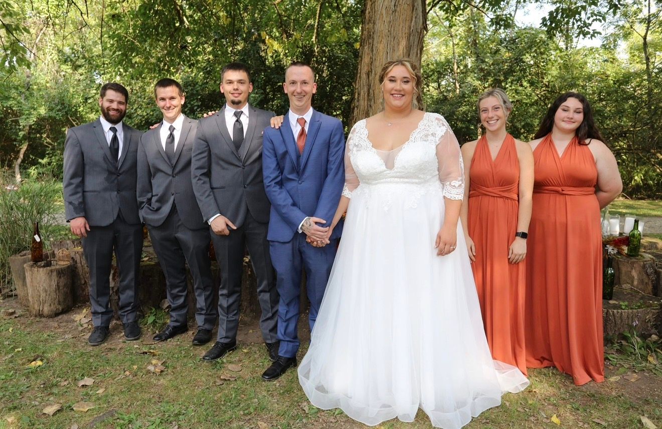 Wedding party posing outdoors with bride in white gown and bridesmaids in rust orange dresses beside groomsmen in gray suits.