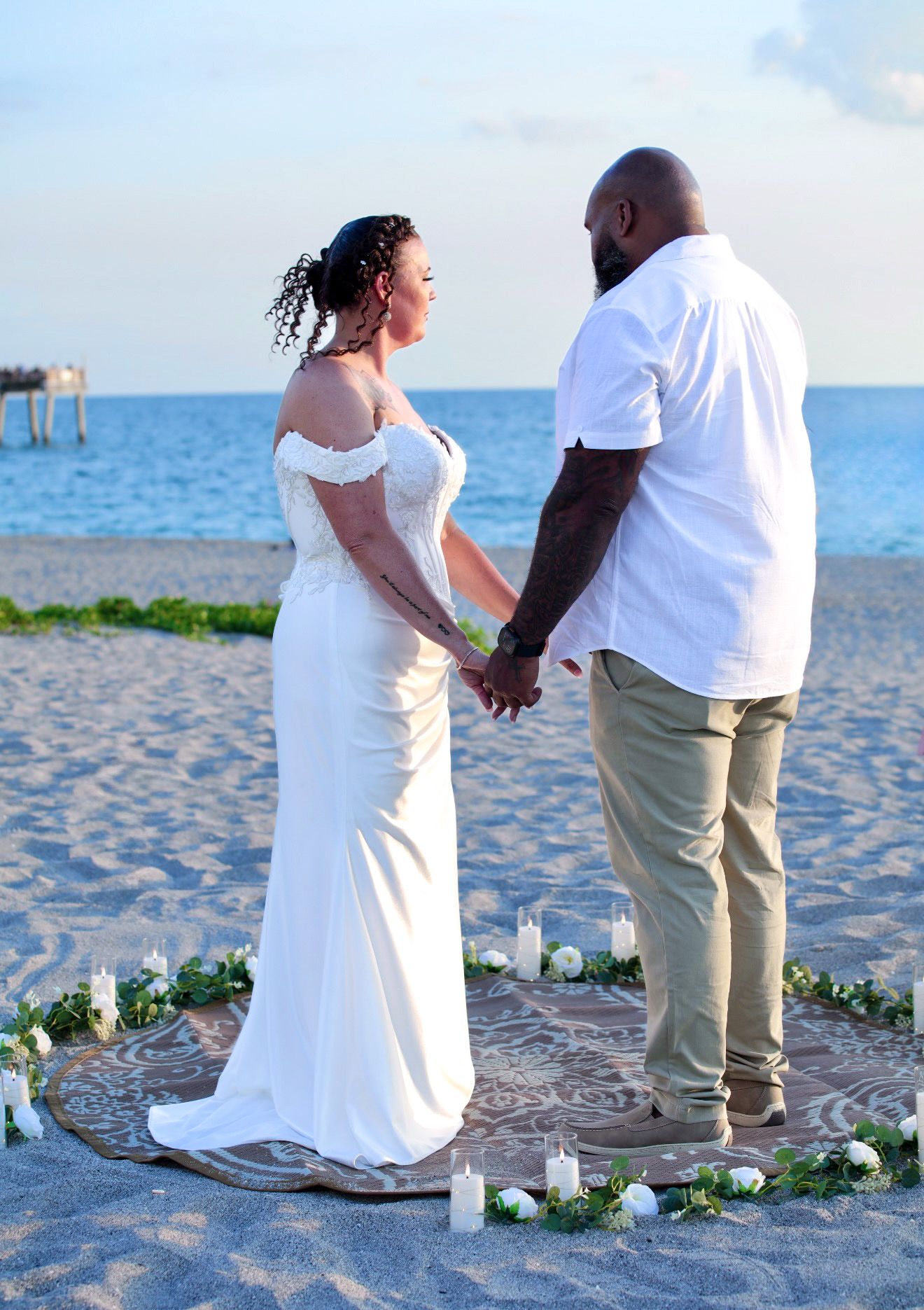 Couple holding hands at a romantic beach wedding ceremony with candles and white roses at sunset.