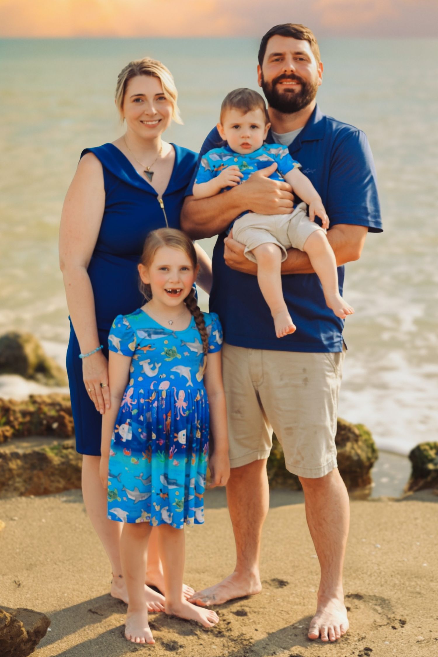 Family of four poses on sandy beach at sunset, parents in blue outfits with young daughter and baby boy.