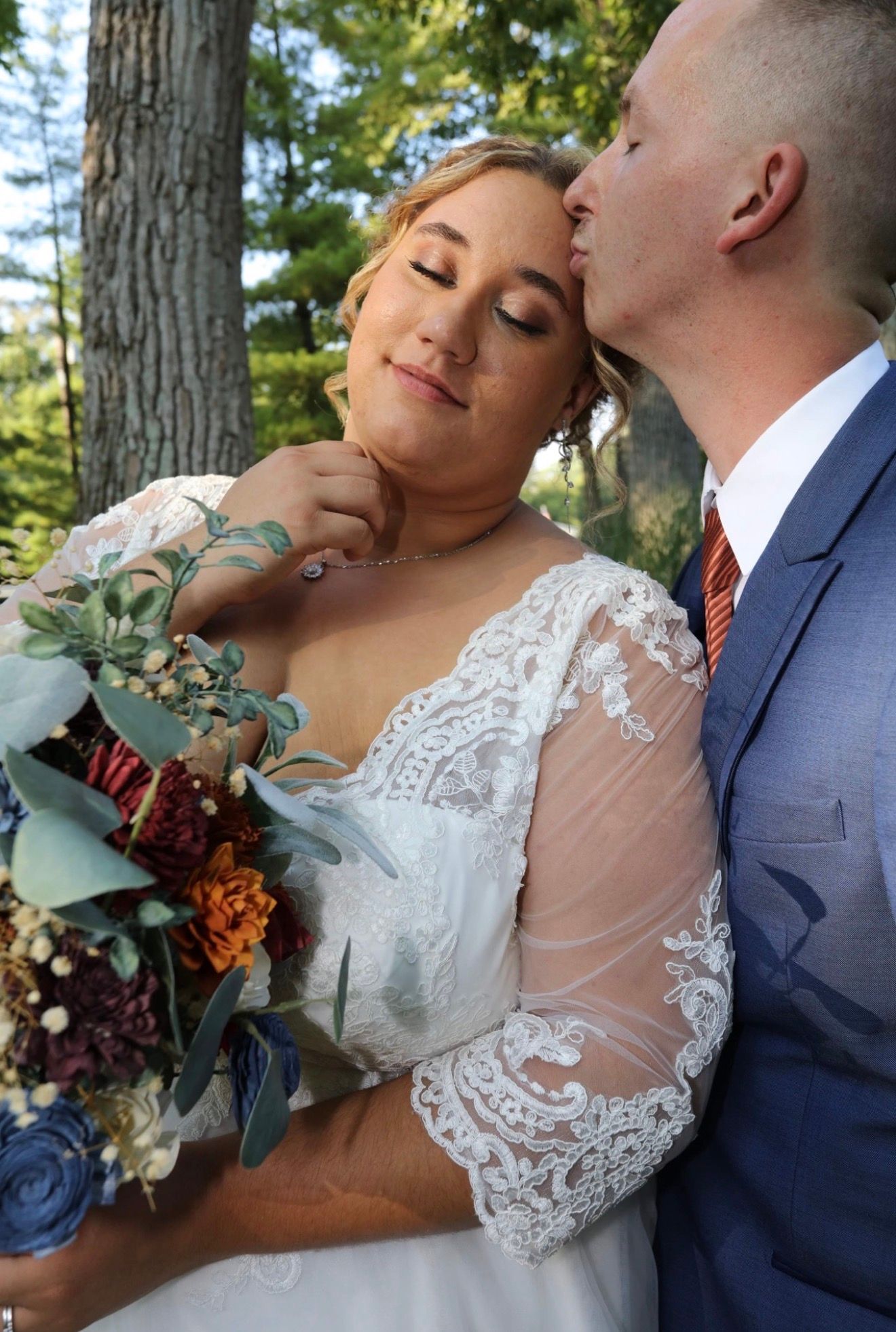 Groom in blue suit kisses bride on cheek as she holds colorful fall bouquet outdoors among trees.