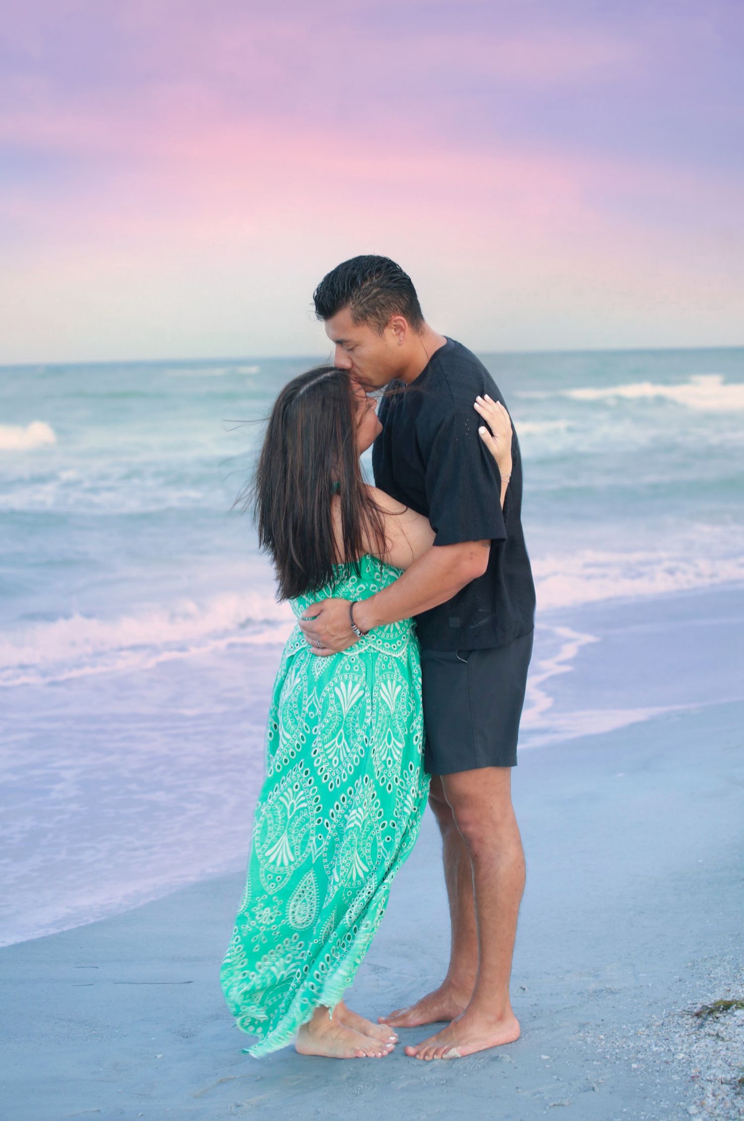 Couple kissing on beach at sunset, woman in green dress, man in black outfit.