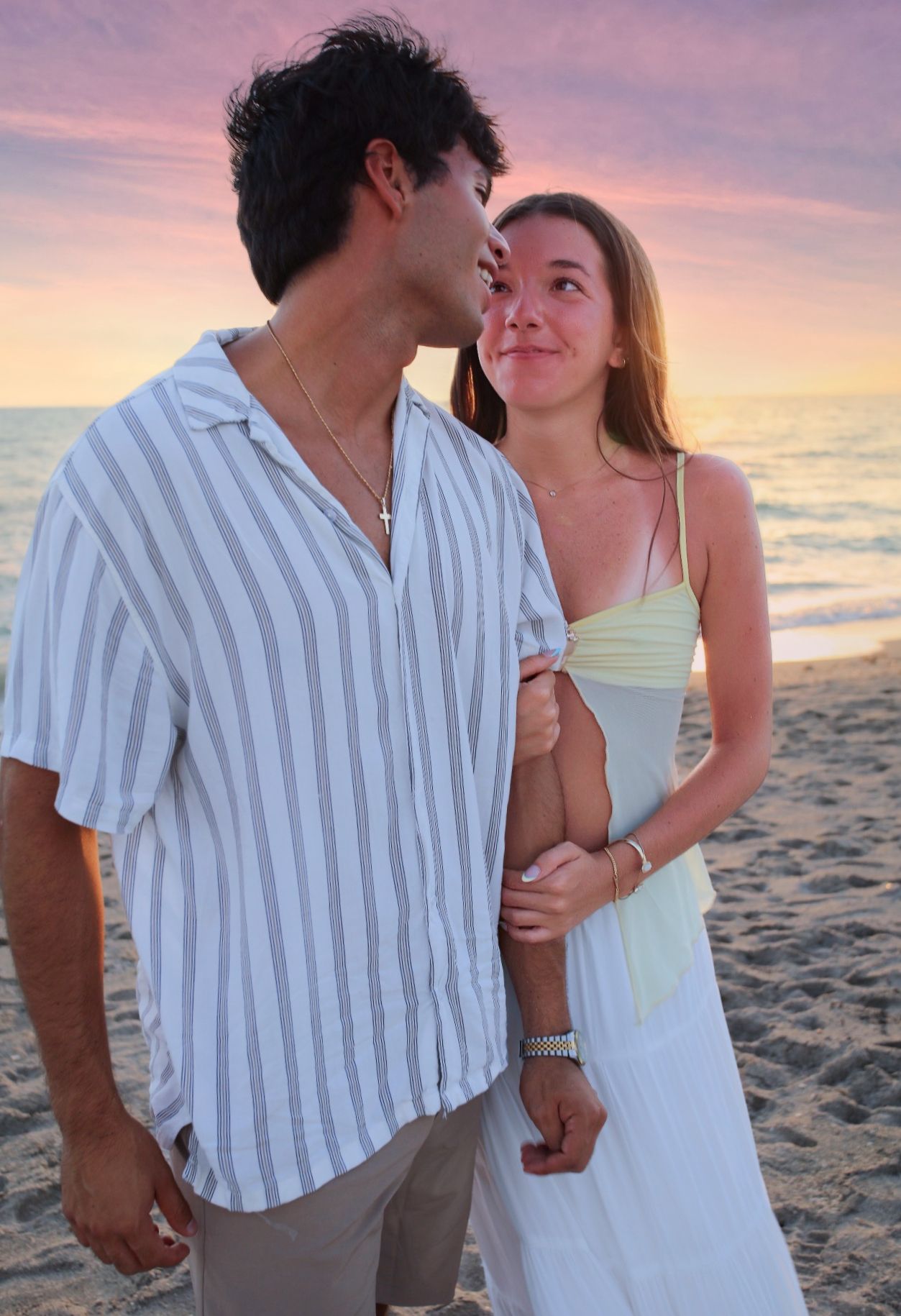 Young couple embracing on a sandy beach during a colorful sunset, wearing casual summer outfits.