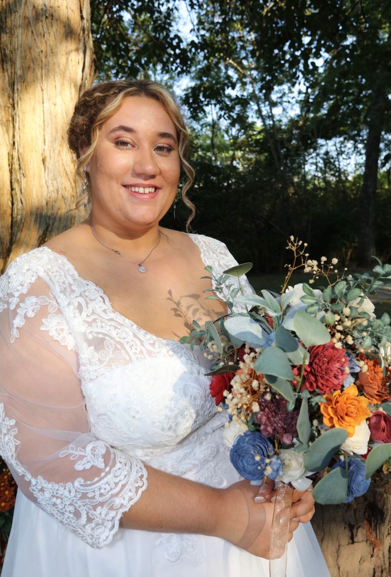 Smiling bride in white lace gown holding colorful bouquet with eucalyptus, rust, and blue flowers outdoors.