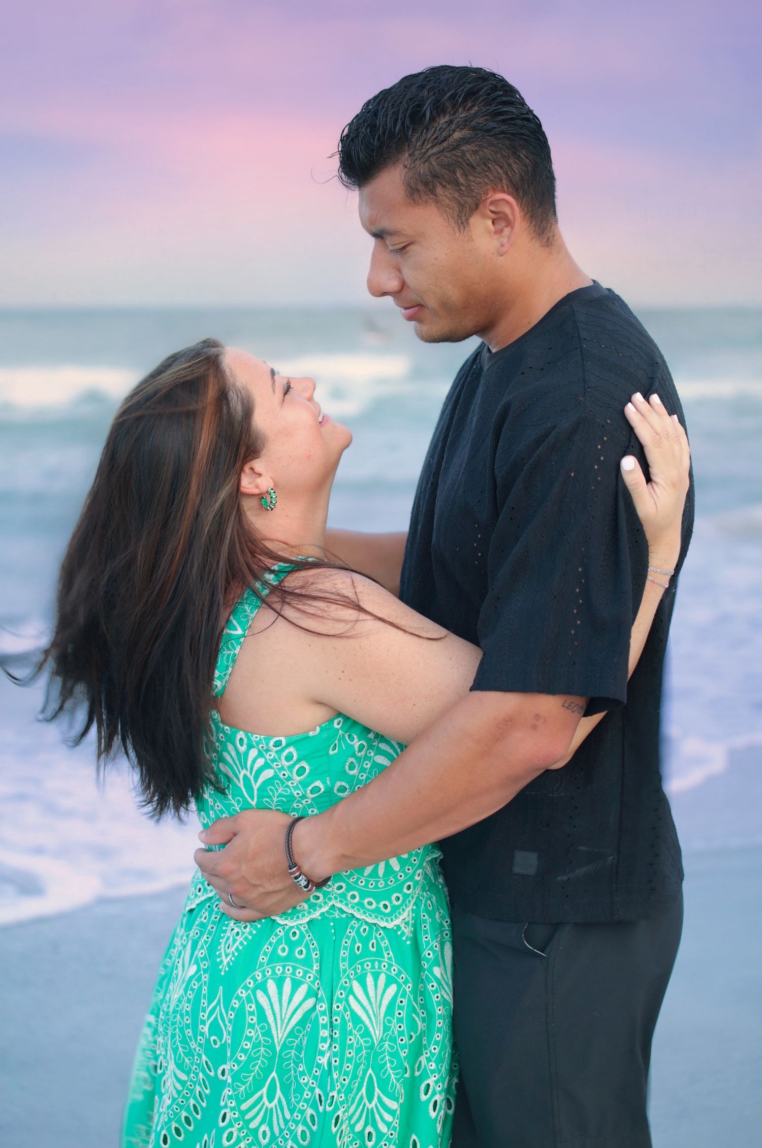 Couple embracing romantically on a beach at sunset, woman in teal dress laughing up at man in black shirt.
