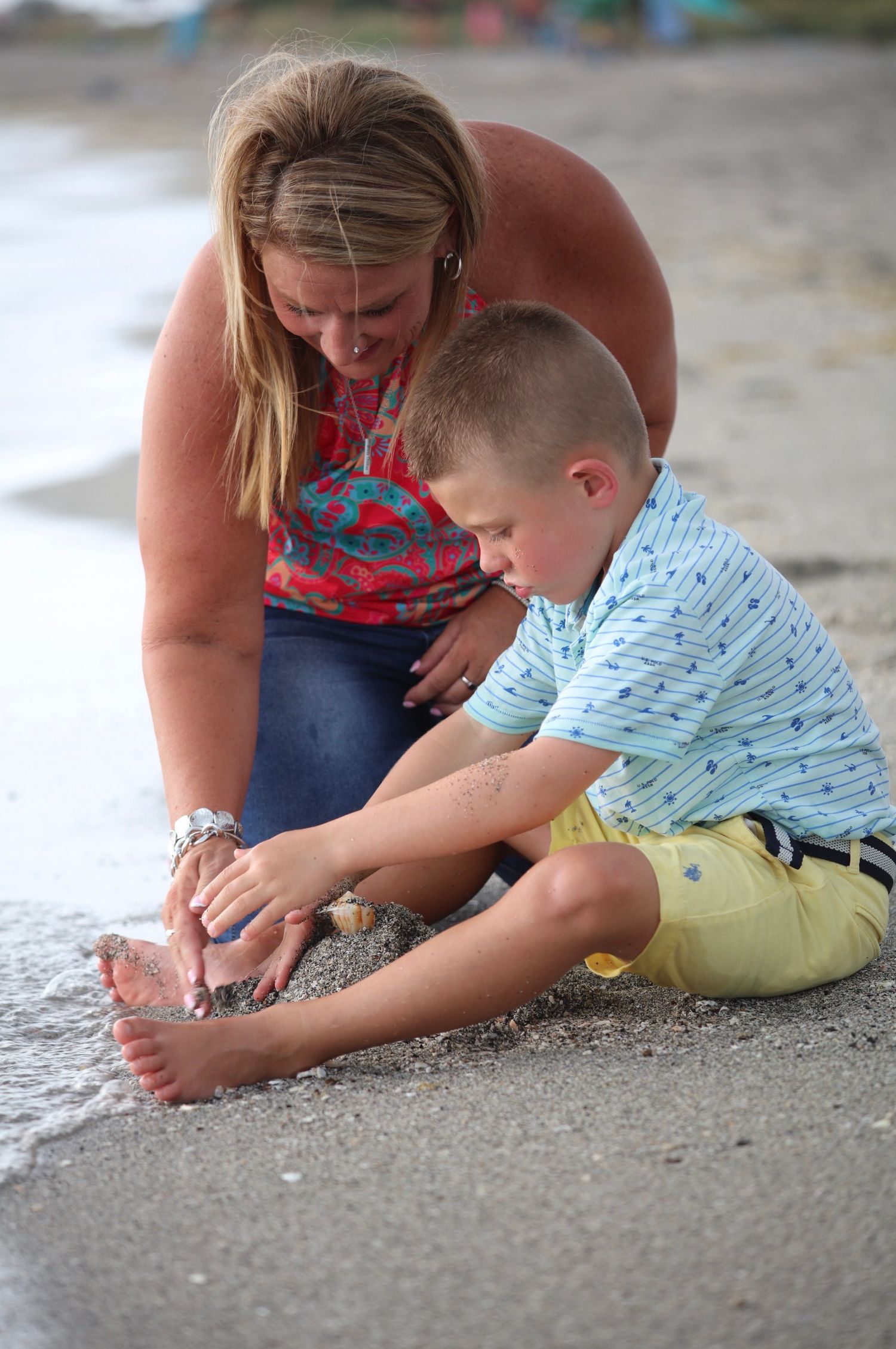 A woman and young boy explore shells together on a sandy beach shoreline.