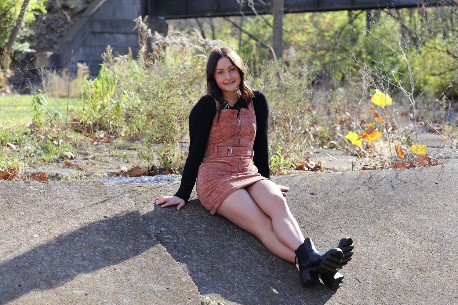 Young woman in pink corduroy dress and black boots sitting outdoors on concrete near a bridge.