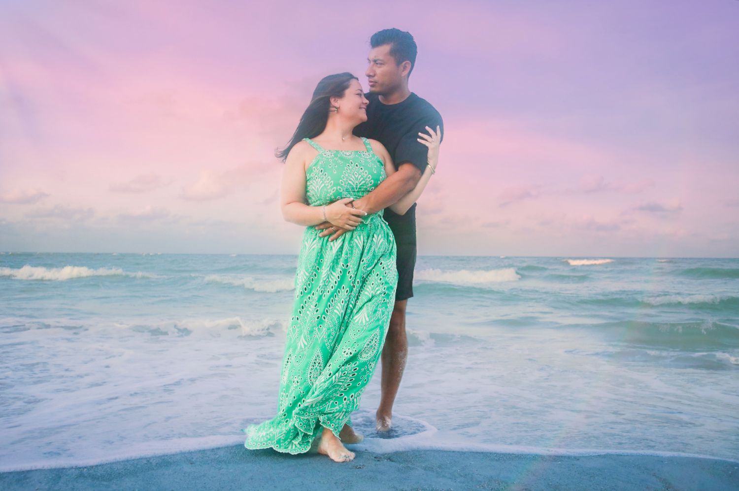 Couple embracing romantically on a beach at sunset, woman in flowing green dress, colorful pink and blue sky backdrop.