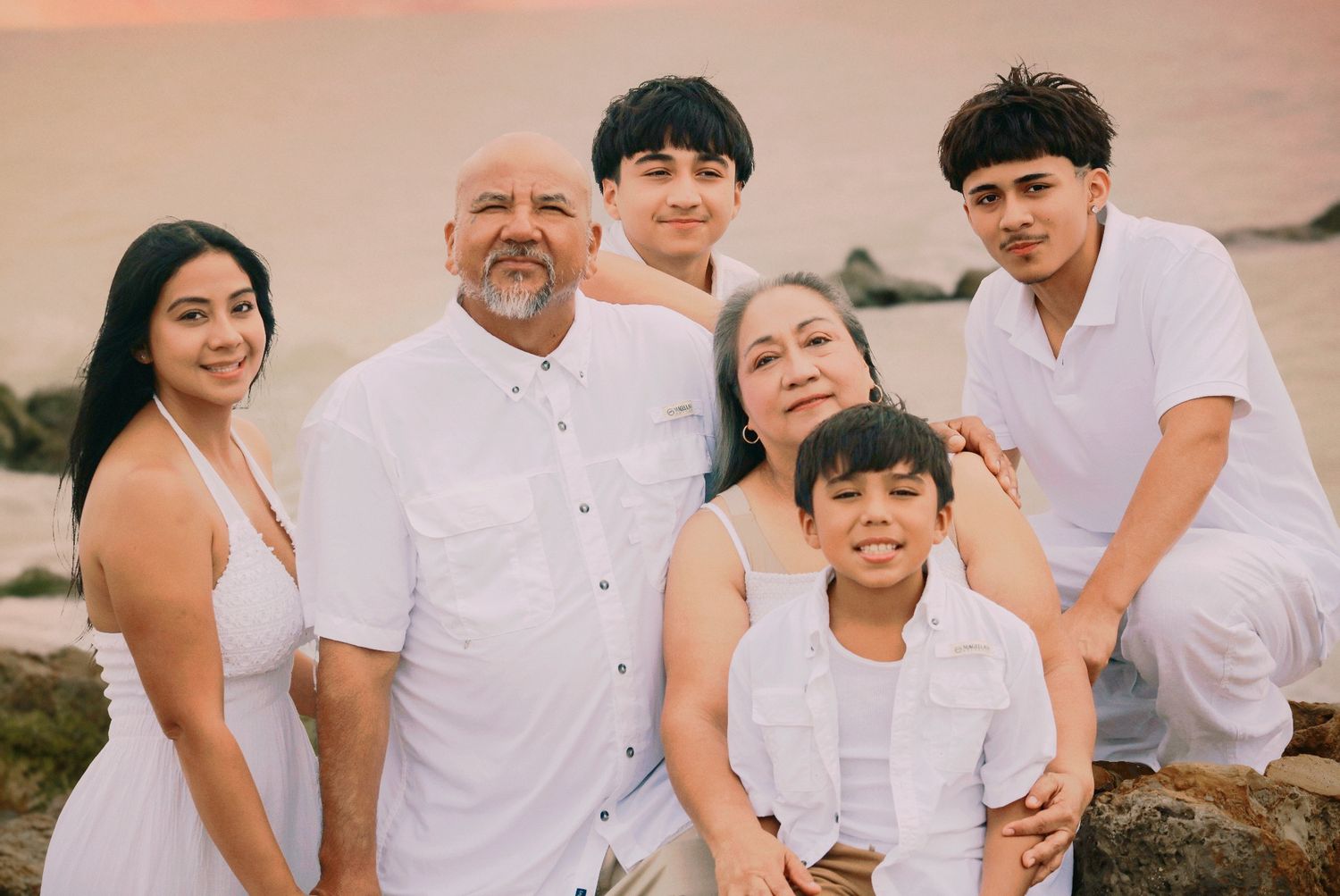 Family of six in white outfits posing together on rocky beach at sunset.