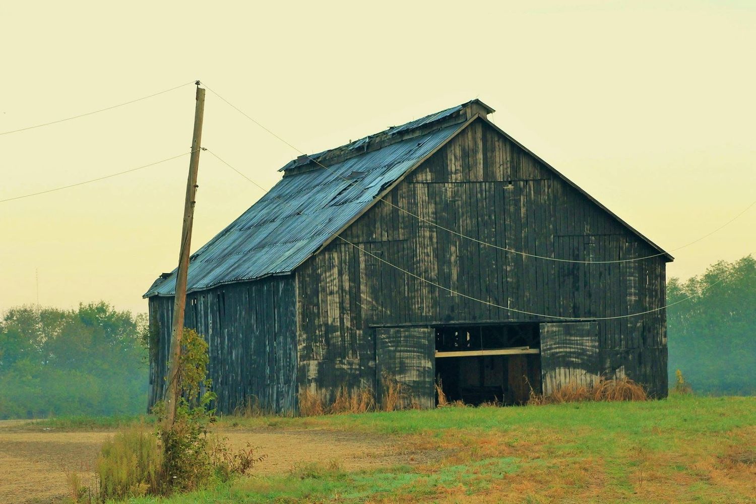 An old weathered wooden barn stands in a rural field under a hazy sky with trees in the background.