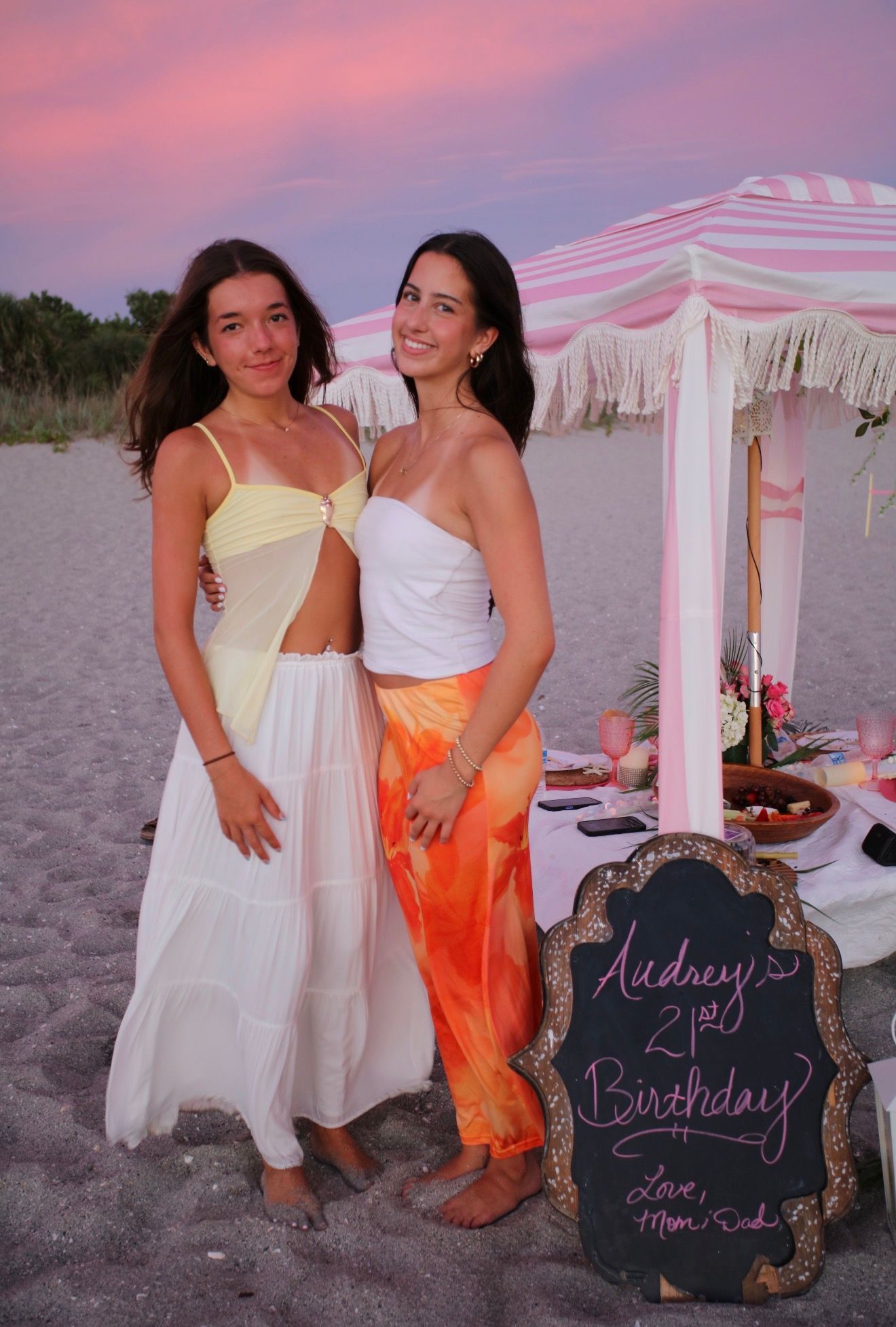Two women pose on a beach at sunset beside a pink striped cabana and a chalkboard sign for Audrey's 21st birthday.