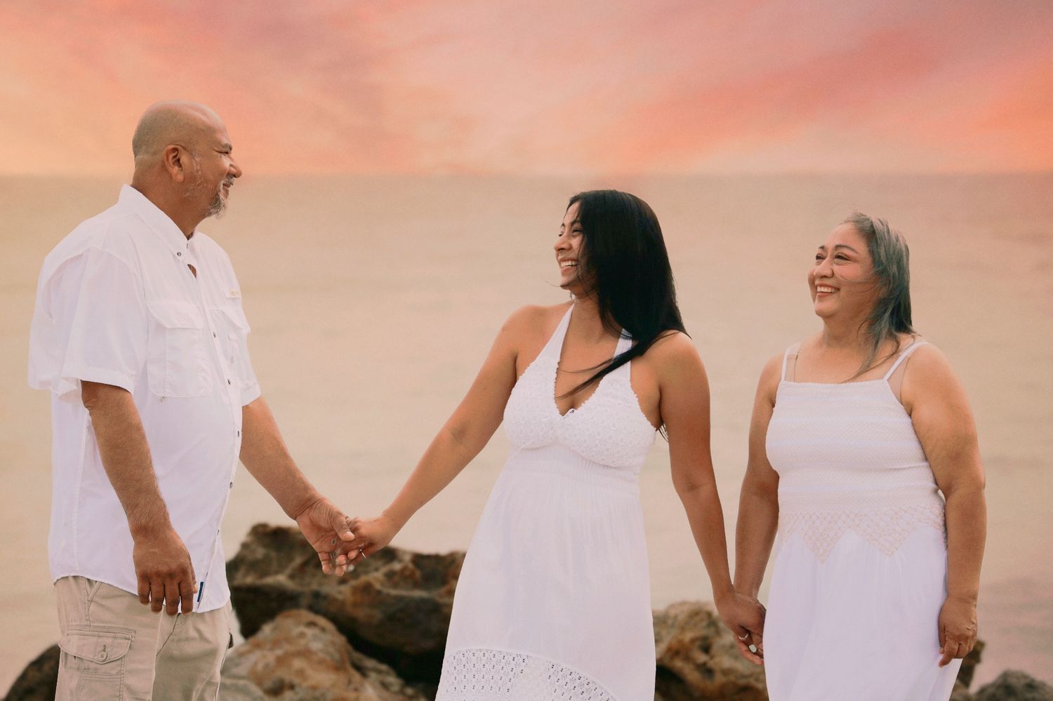 Family of three in white outfits holding hands on a beach at sunset.