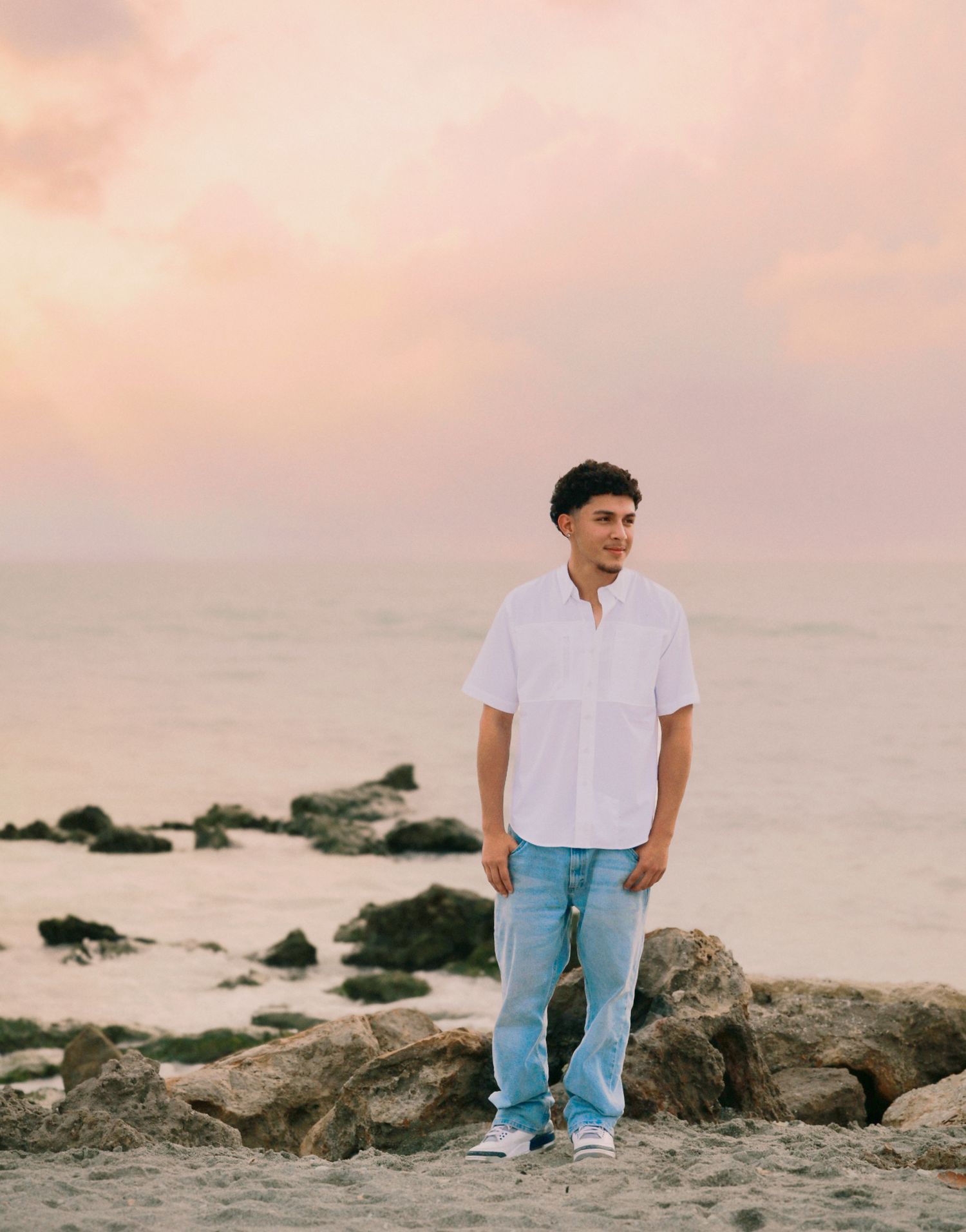 Young man in white shirt and jeans standing on rocks at beach during pink sunset.