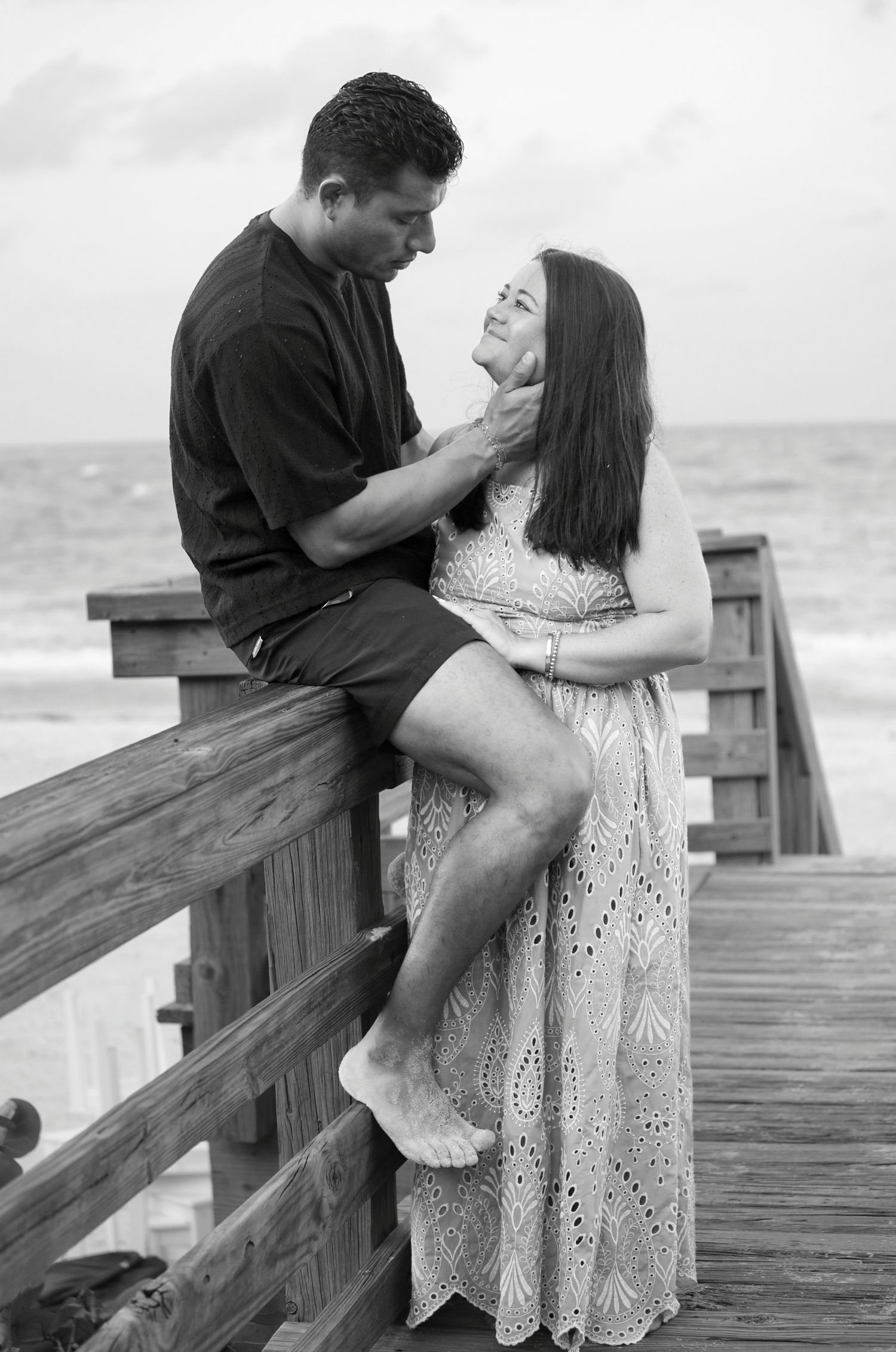 Black and white romantic couple on wooden boardwalk by ocean, man sitting on railing holding womans face.