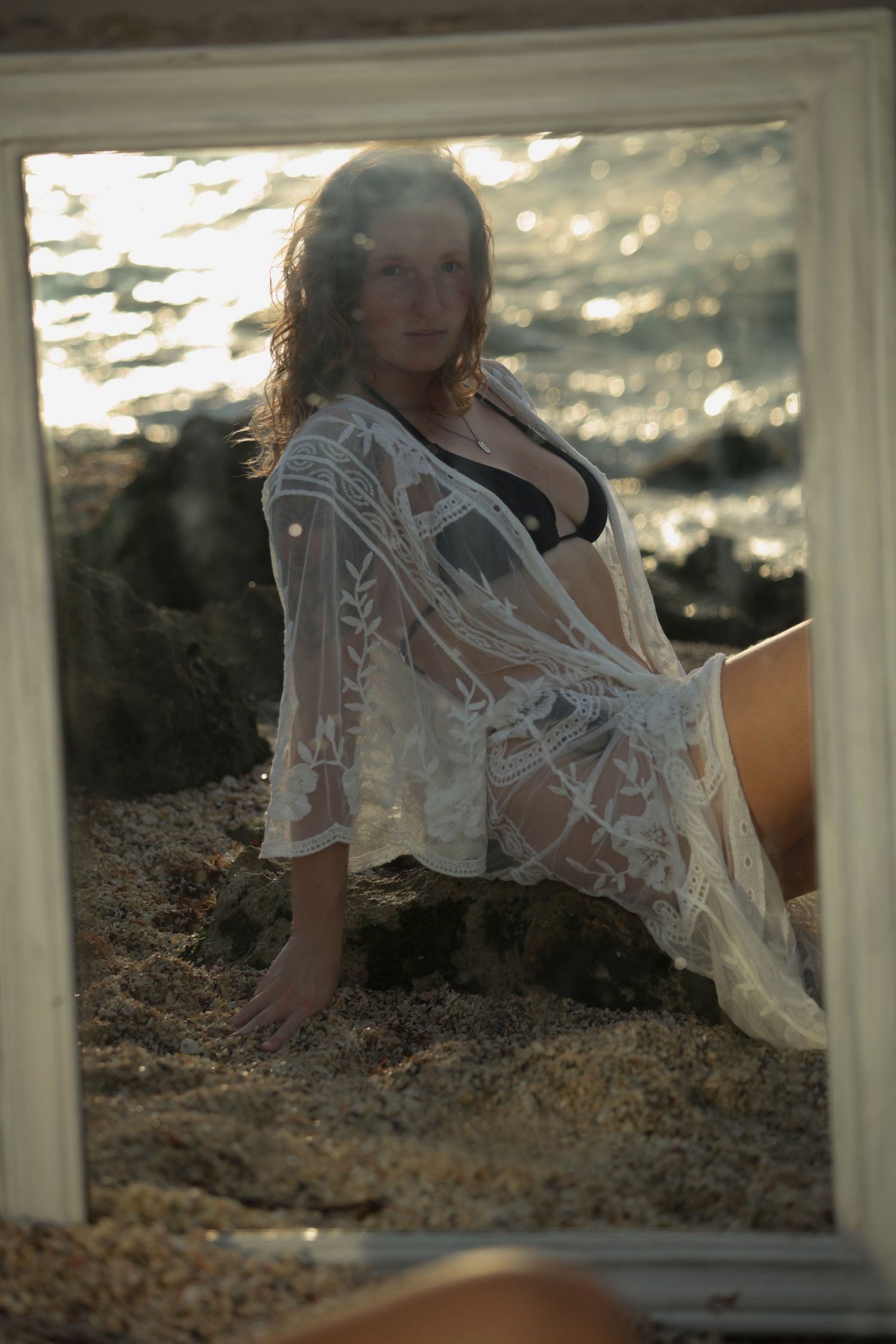 Woman in black bikini and white lace cover-up sitting on sandy beach rocks viewed through a white picture frame mirror.