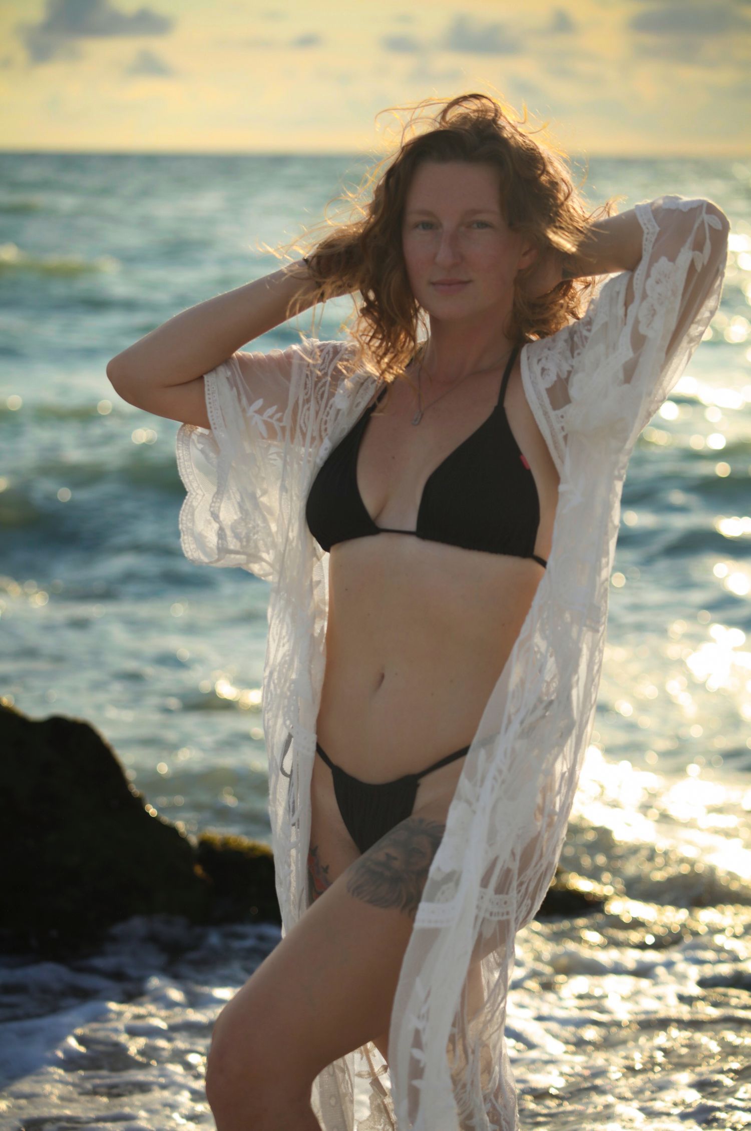 Curly-haired woman in black bikini and white lace robe poses at sunny beach shoreline.