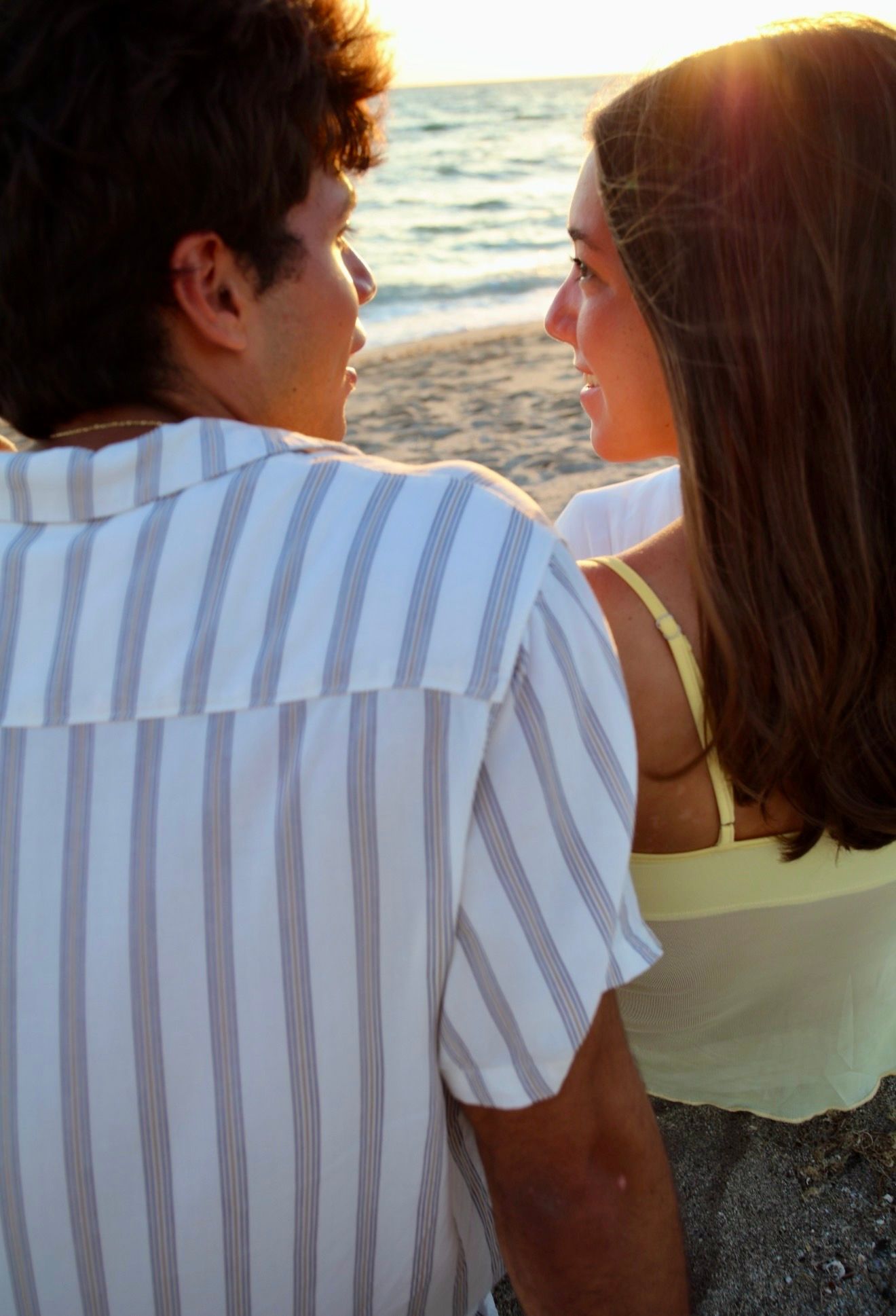 A couple shares a romantic moment on a sunlit beach, smiling at each other near the water.