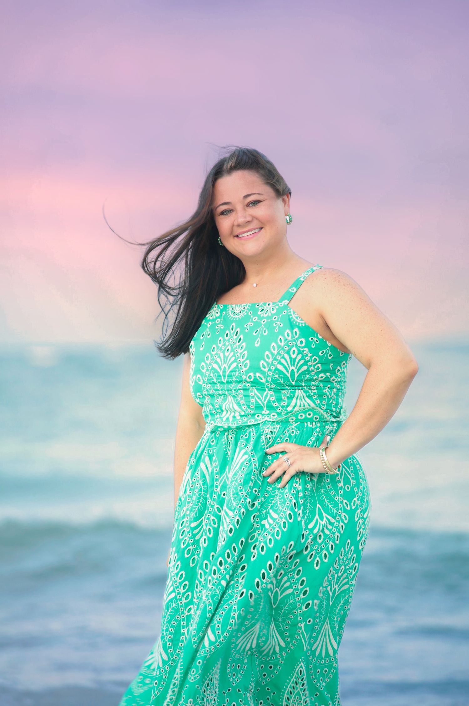 Woman in green maxi dress smiling on beach at sunset with wind blowing hair.