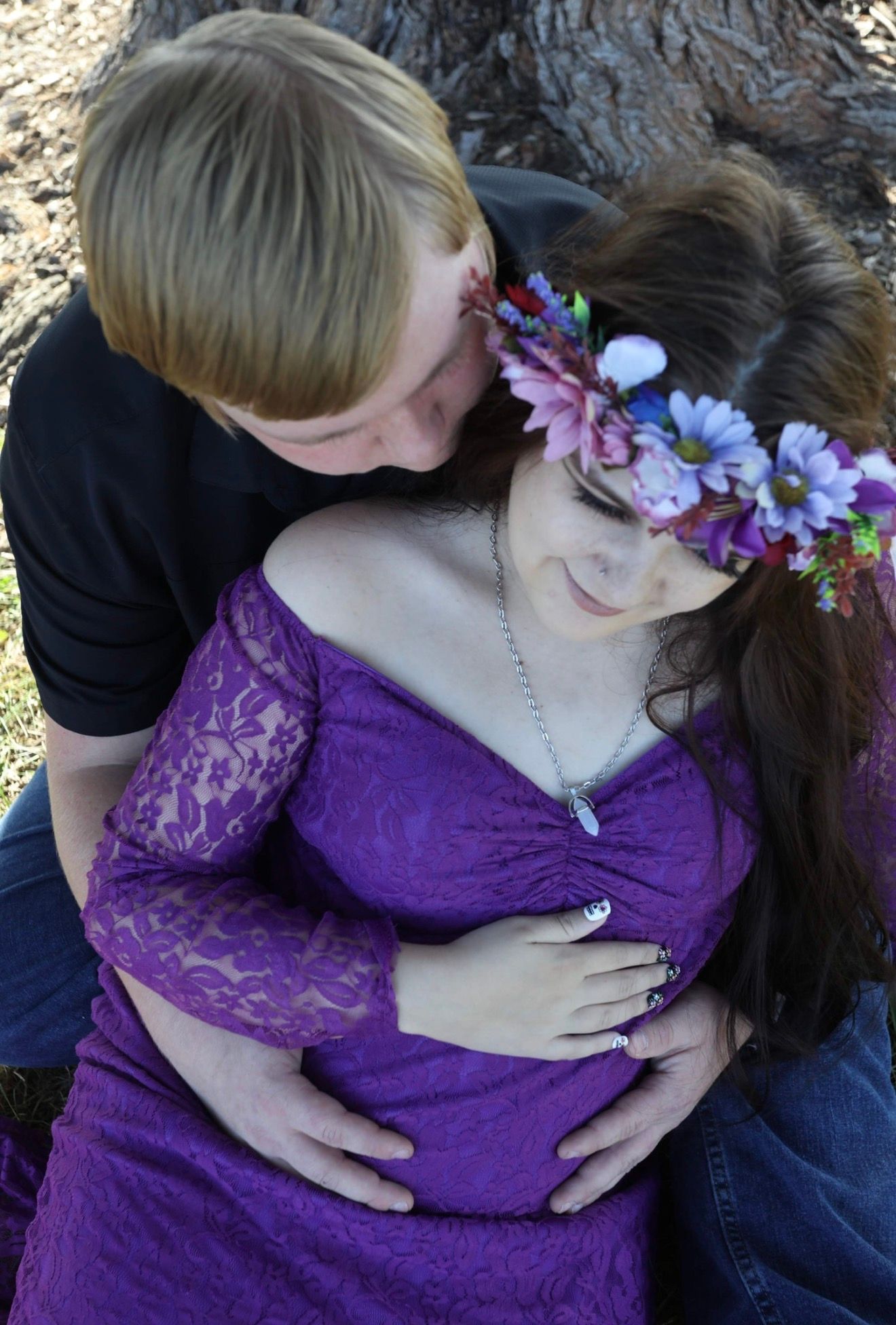 Couple sharing a tender maternity photo outdoors, woman in purple lace dress with floral crown, partner kissing her head.
