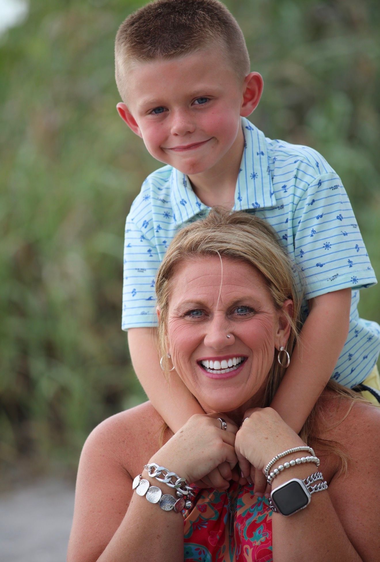 A smiling woman and young boy in blue polo shirt pose together outdoors, the boy resting his chin on her head.