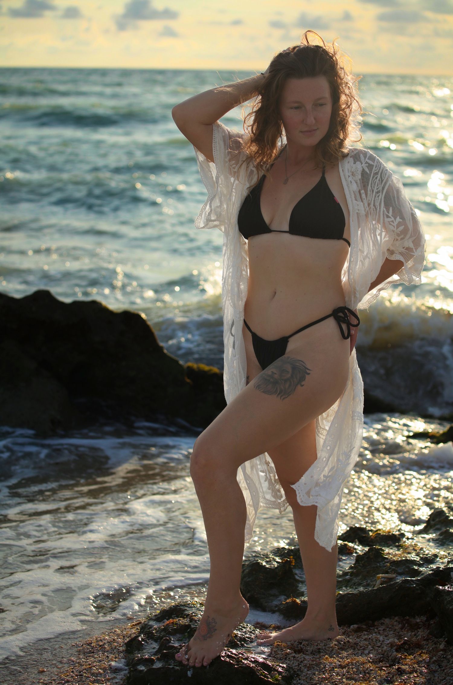Woman in black bikini and sheer white lace cover-up posing on rocky shoreline with ocean waves at golden hour.