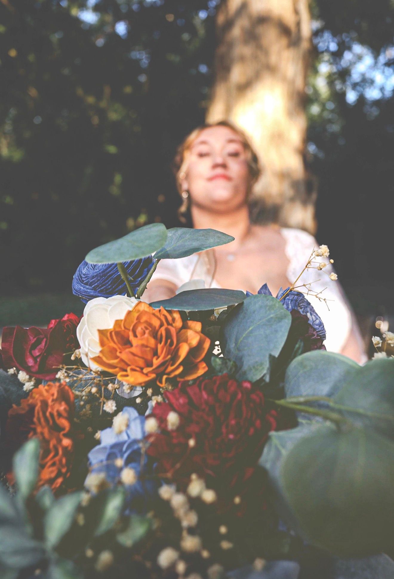 Bride in white dress holds colorful bouquet with orange, red, and blue flowers surrounded by lush greenery outdoors.