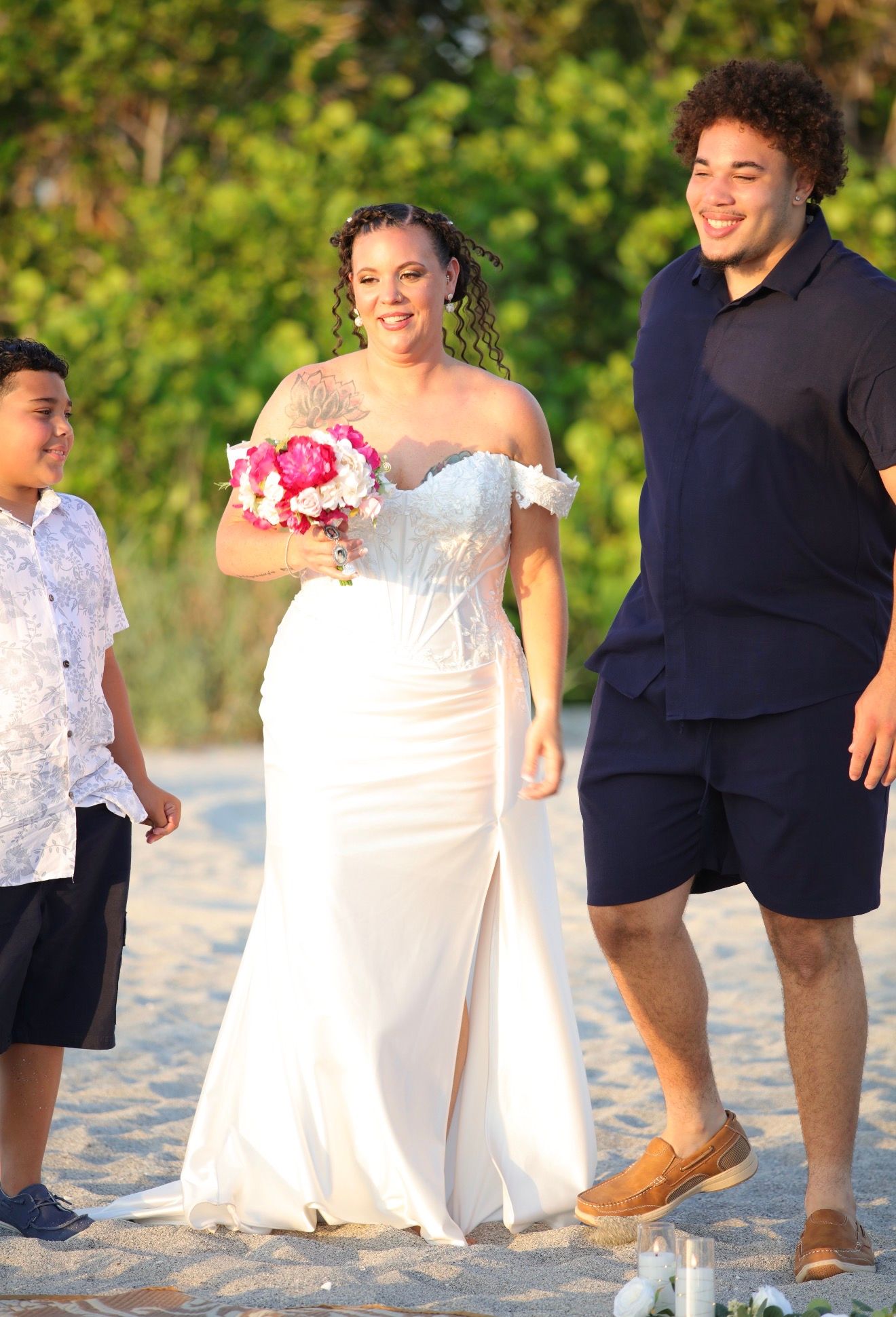 Bride in white off-shoulder gown holds pink bouquet walking on beach with two guests at sunset wedding.