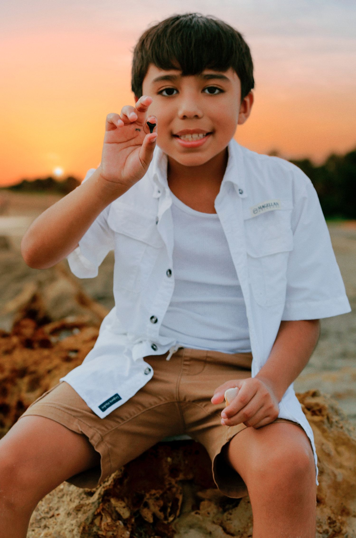 Young boy in white shirt and khaki shorts sitting on rocks at sunset beach, smiling and holding a small object.