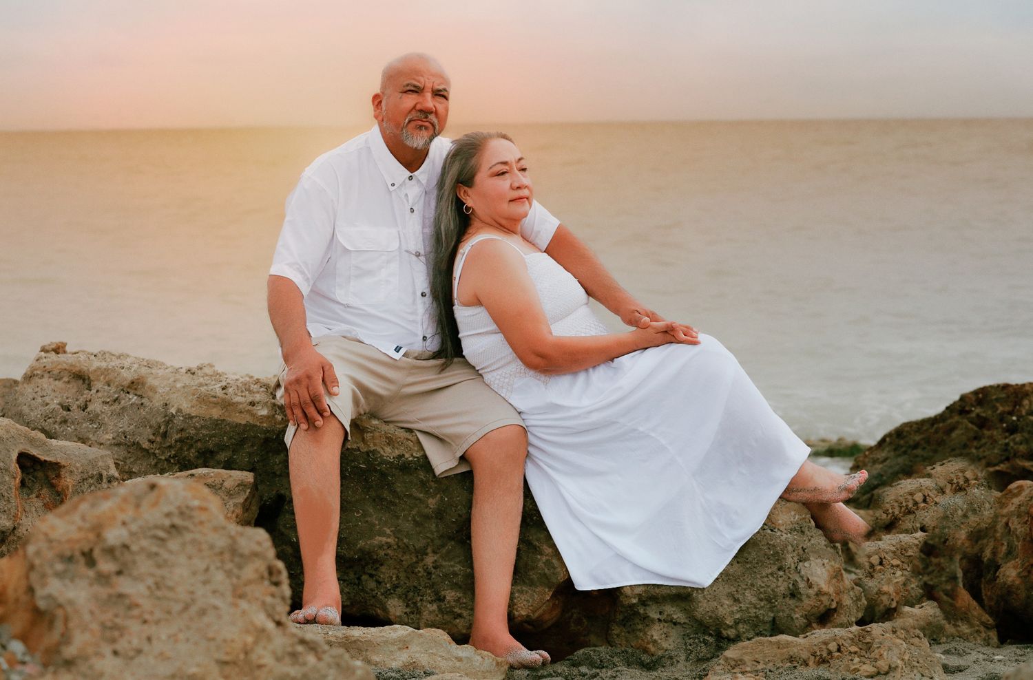 Couple in white outfits sitting on rocks by the ocean during a golden sunset.