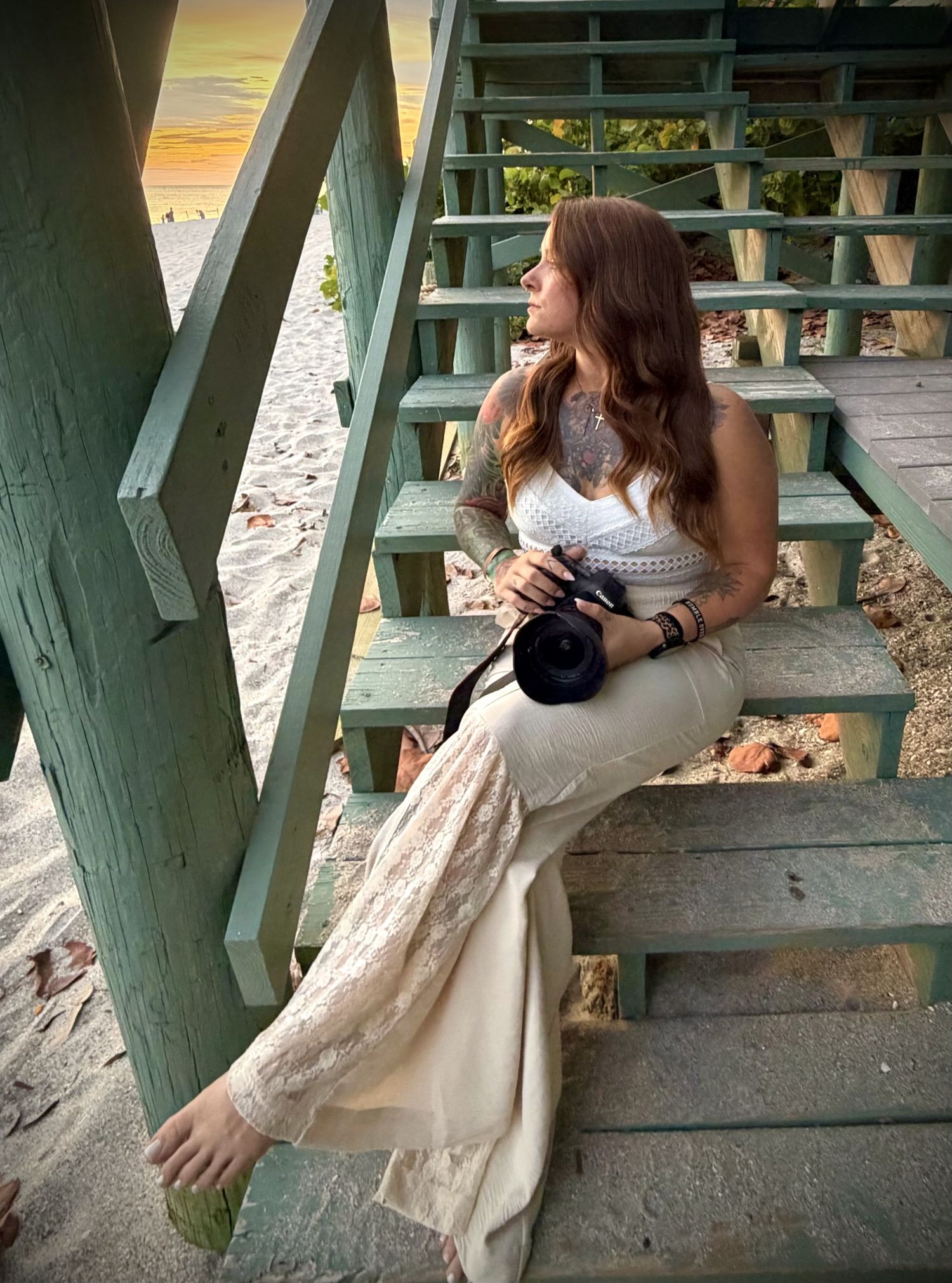 Woman with long hair sits on beach stairs holding a camera, wearing a white top and flowy beige skirt at sunset.