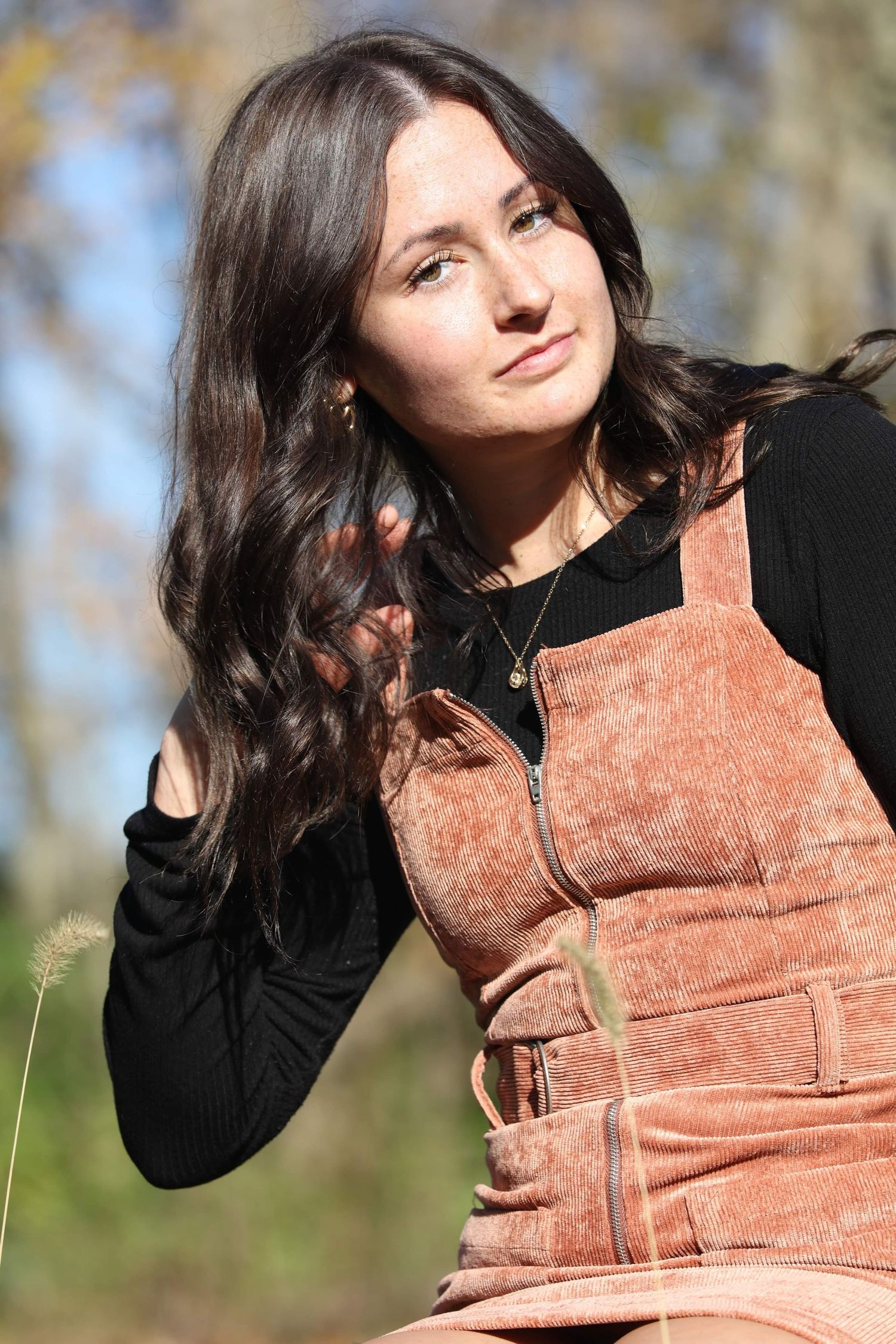 Young woman with long dark hair wearing rust corduroy overalls over black top, posing outdoors in natural light.