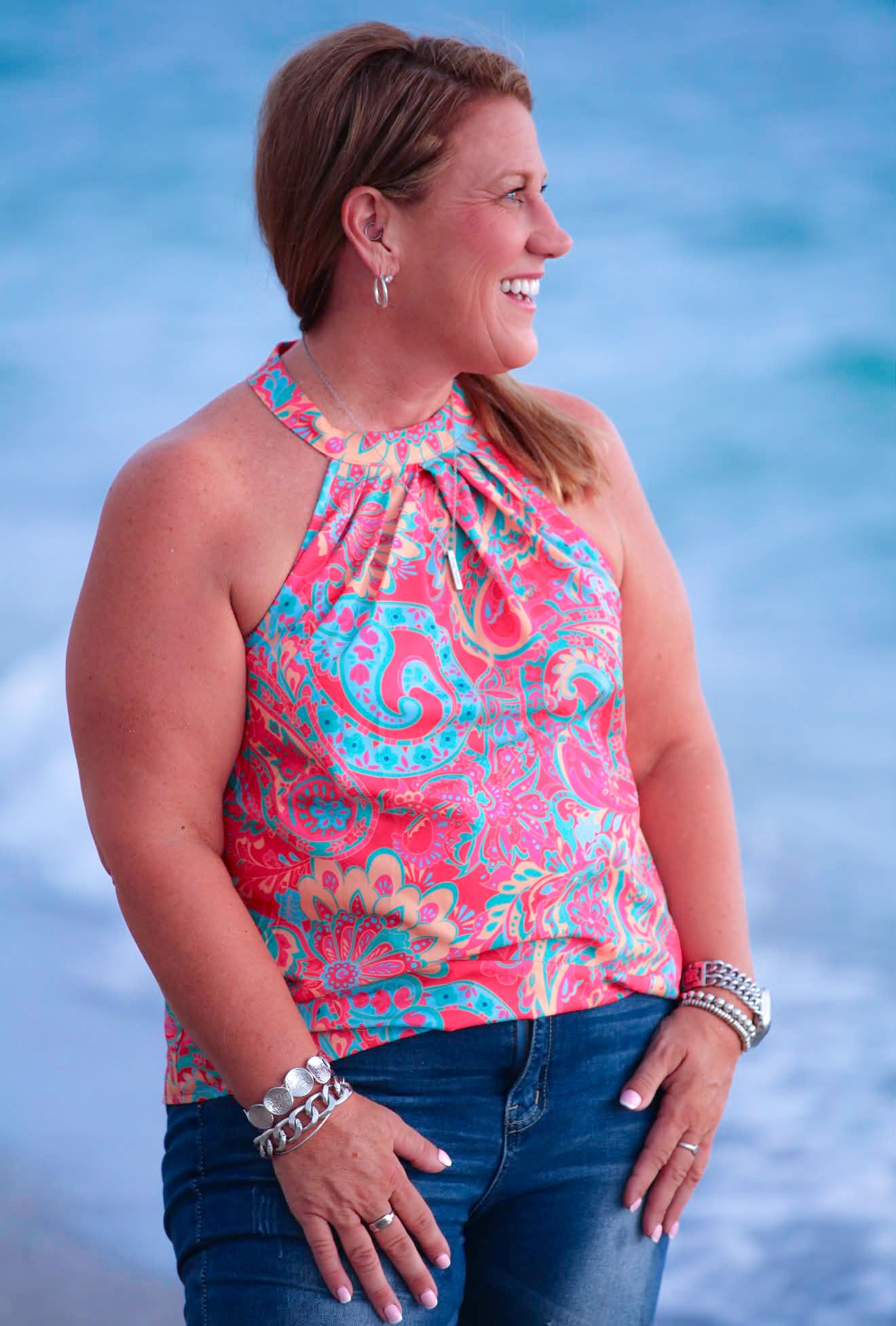Woman in colorful paisley halter top and jeans smiling at the beach with blue ocean in the background.