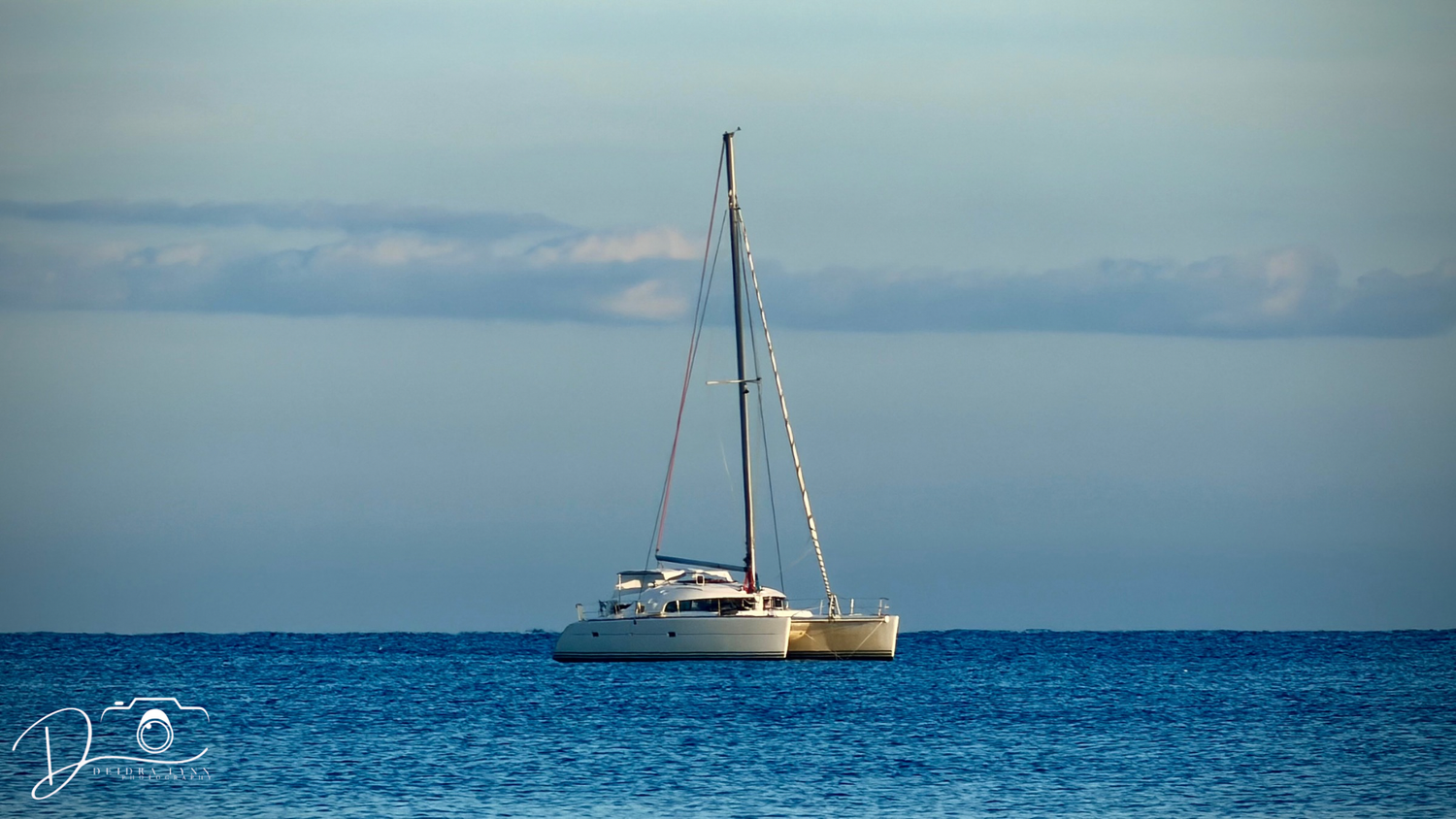 White catamaran sailboat anchored on calm blue ocean waters under a partly cloudy sky.