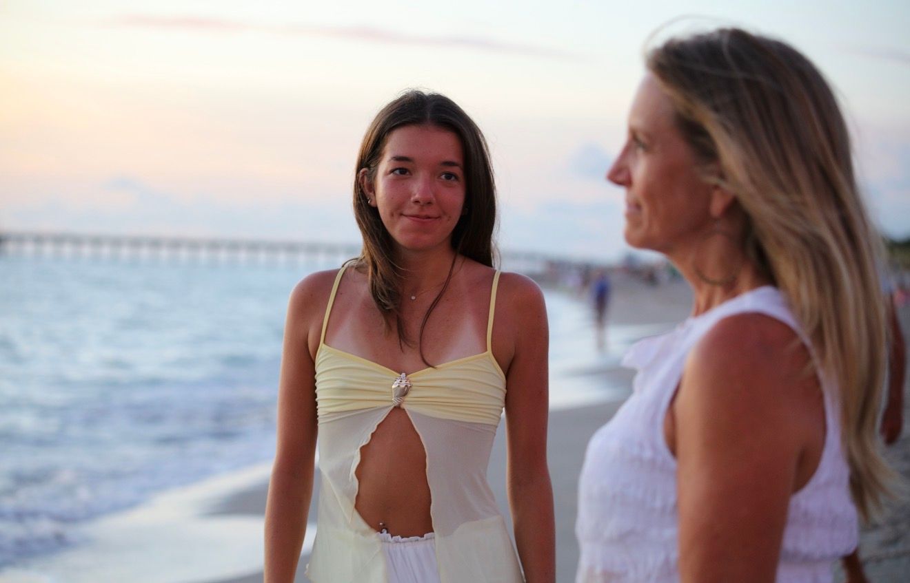 Two women in white outfits stand on a beach at sunset with a pier visible in the background.