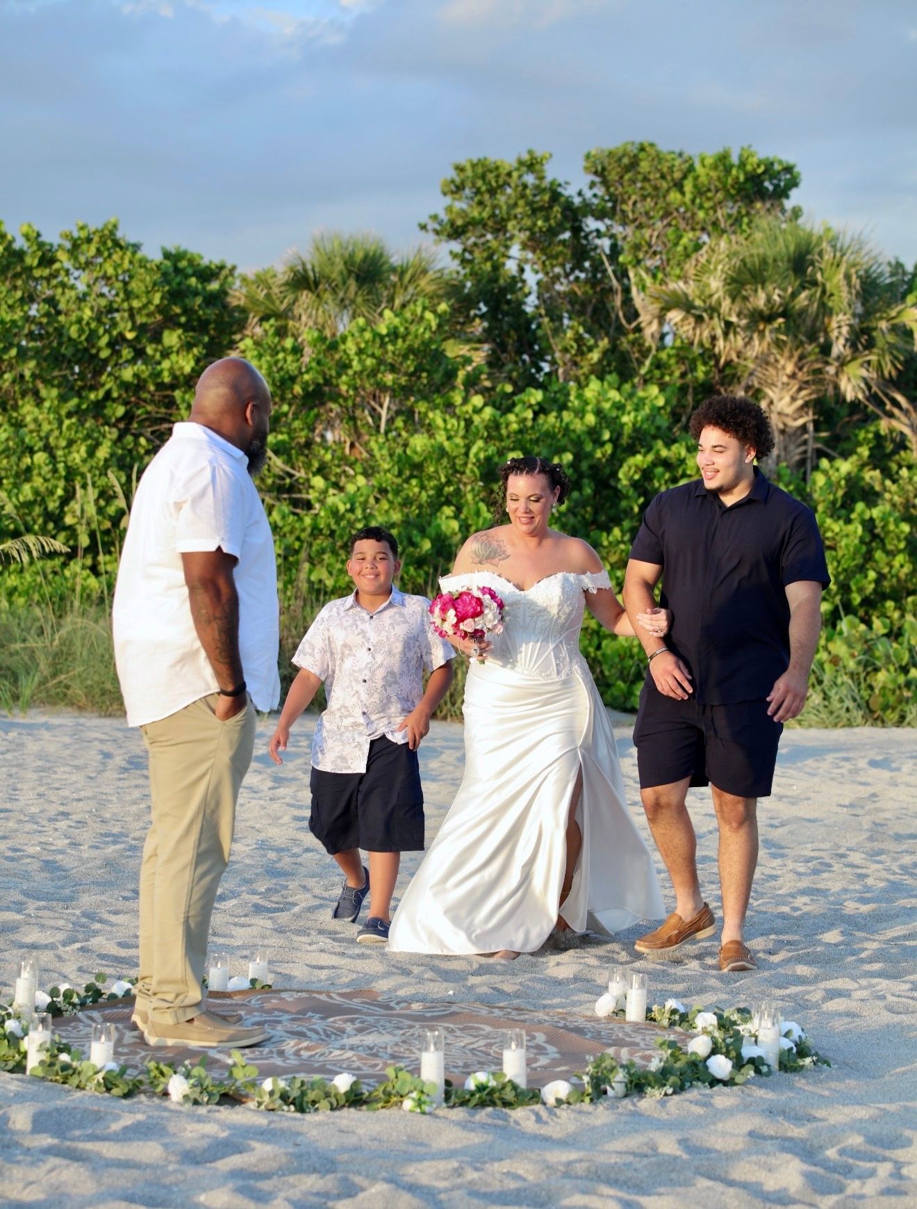 Bride in white gown holds bouquet with family on sandy beach, surrounded by candles and flowers at sunset.