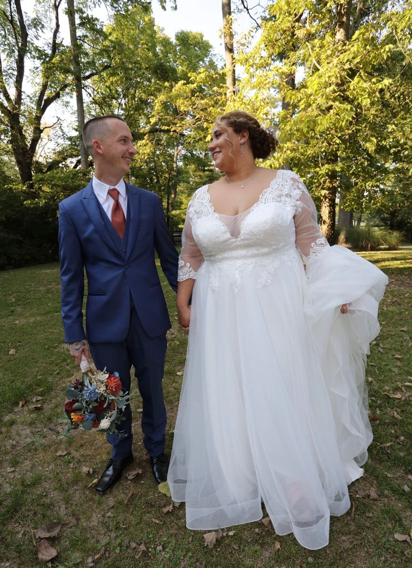 Bride in white lace gown and groom in blue suit hold hands smiling outdoors on their wedding day.
