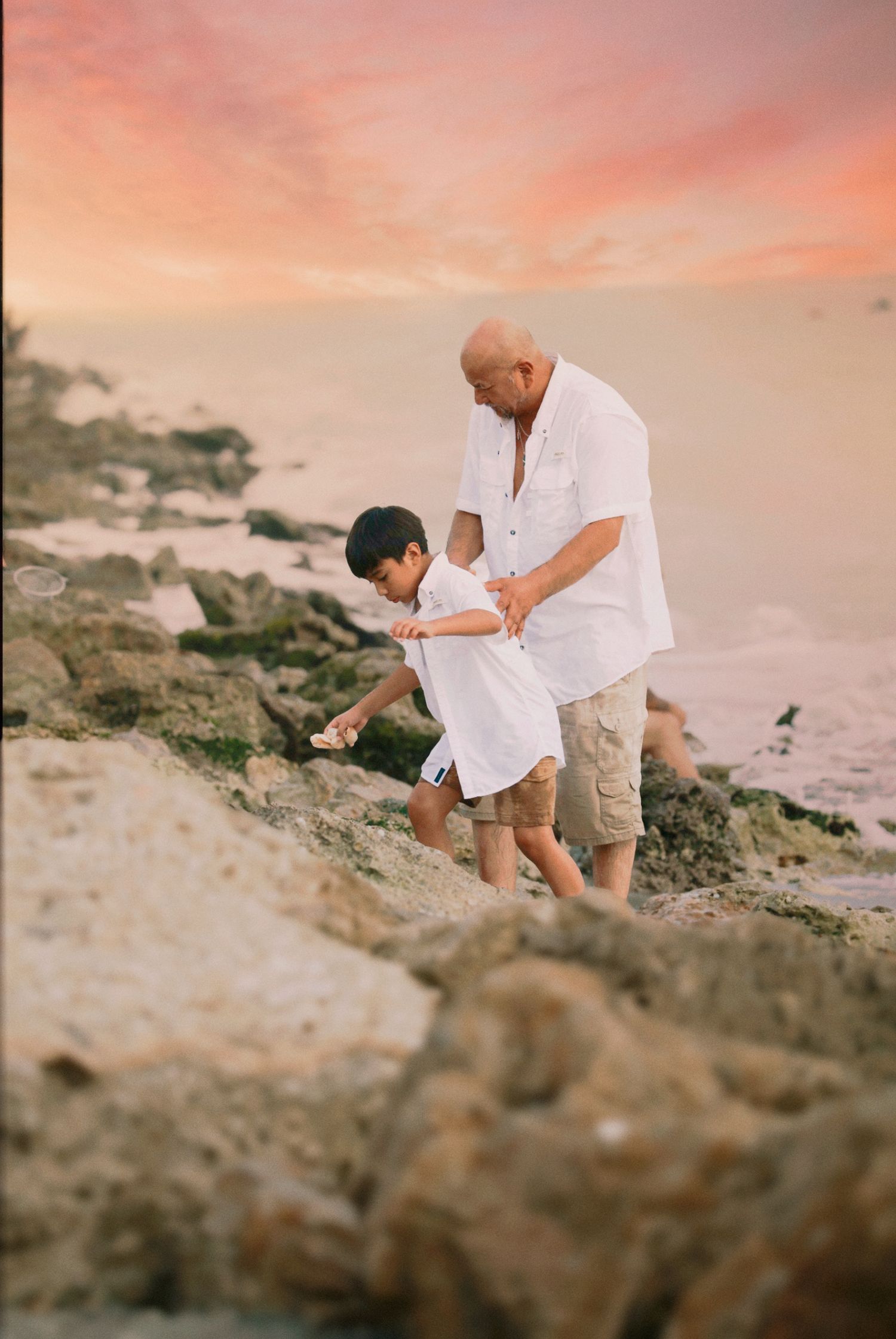 A grandfather and young boy explore rocky shoreline together at sunset, both wearing white shirts near the ocean.