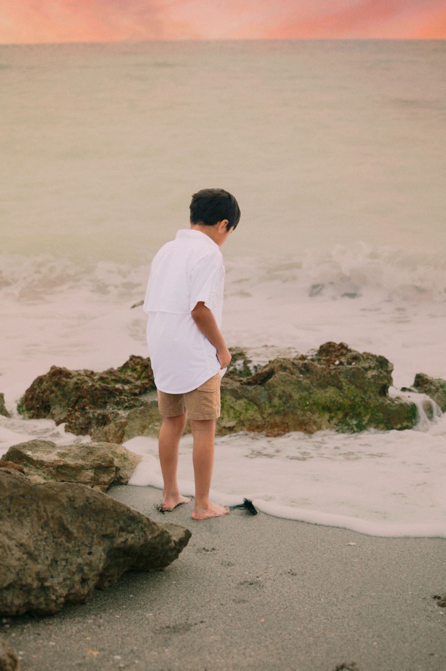 Young boy in white shirt and tan shorts standing barefoot by ocean rocks at sunset.