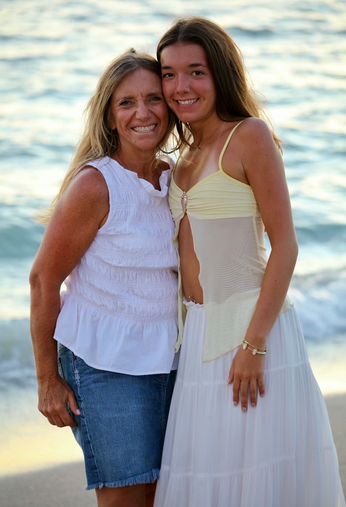Smiling blonde woman and teenage girl posing together on beach at sunset near ocean waves.