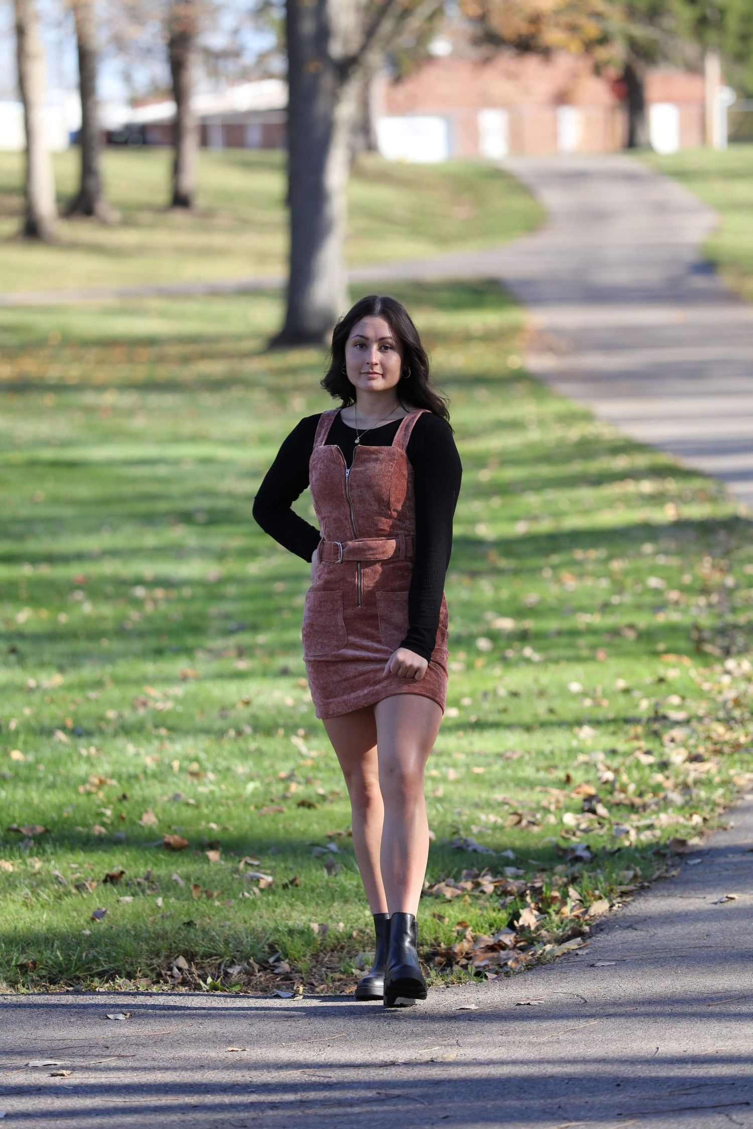 Young woman in brown pinafore dress and black top walking on a park path in autumn.