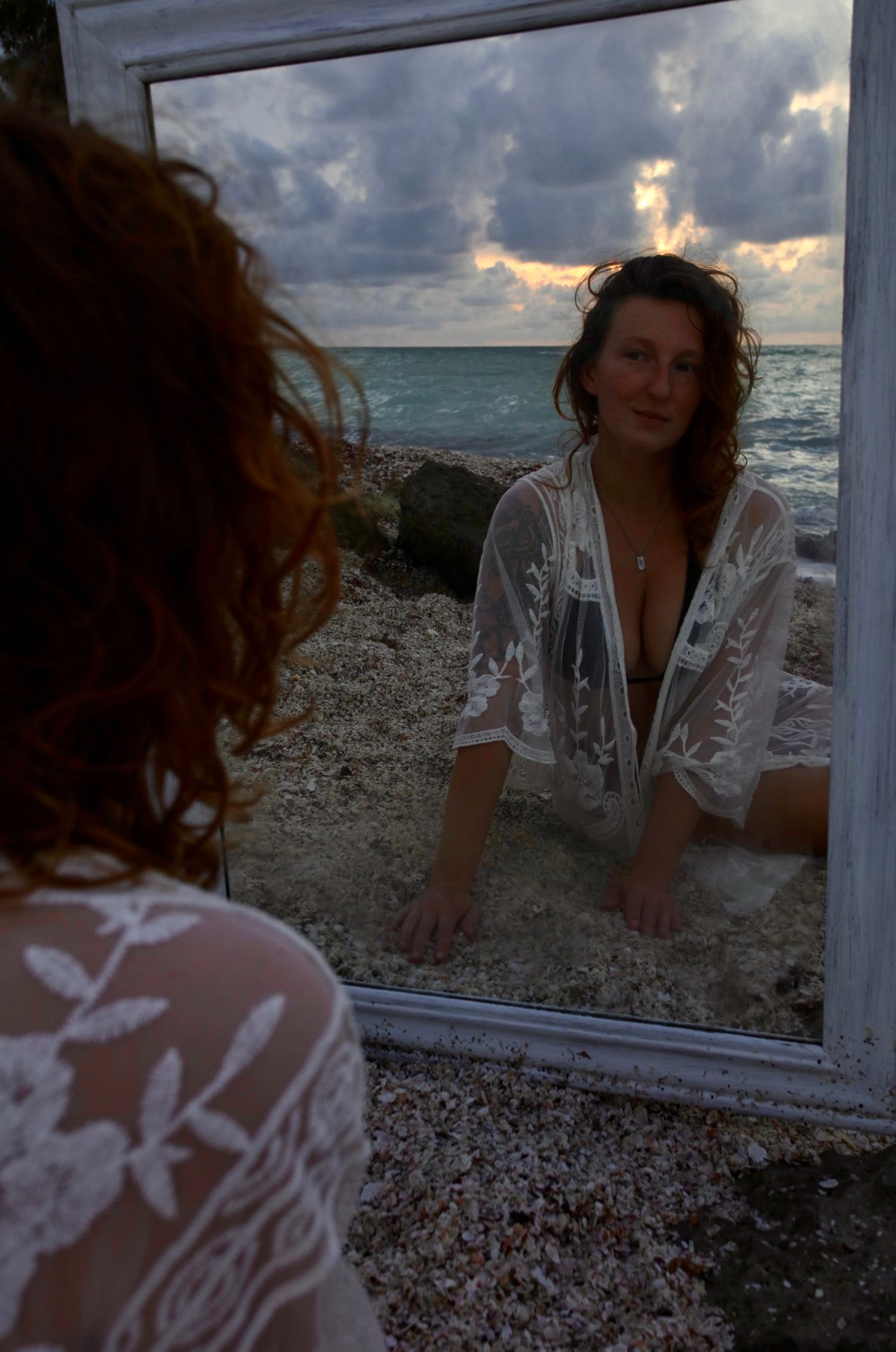 Woman in sheer white lace robe gazing at her reflection in a large mirror on a pebble beach at dusk.
