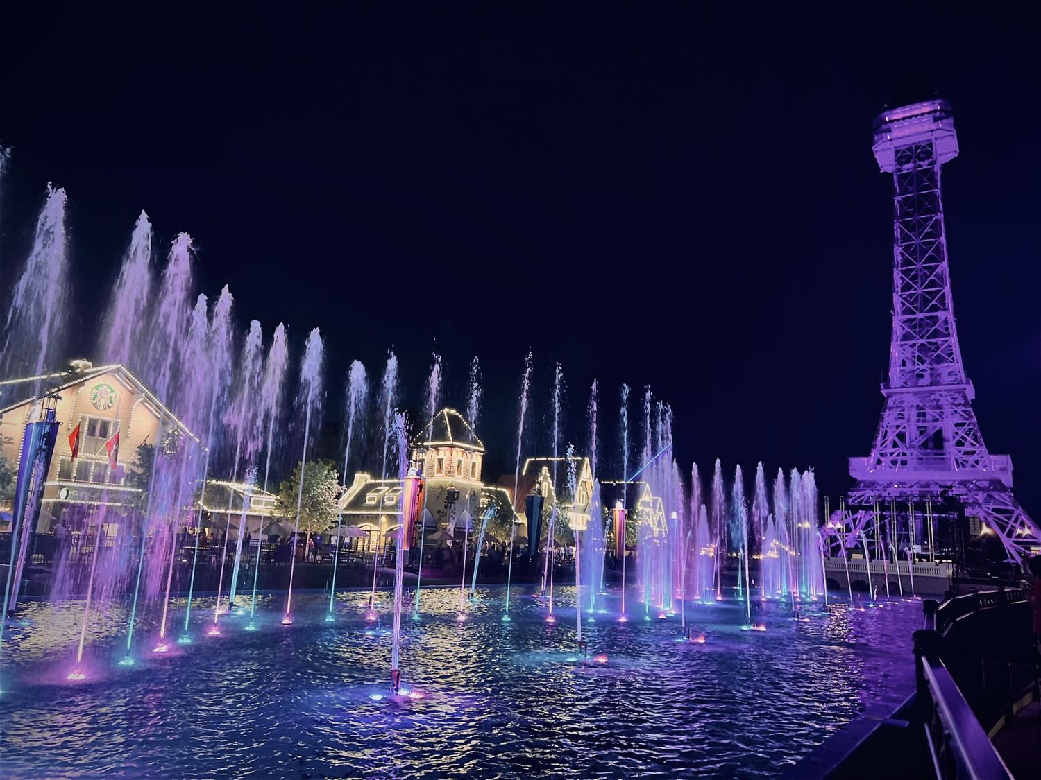 Colorful illuminated fountain show at night with purple and pink water jets near an Eiffel Tower replica at Kings Island.