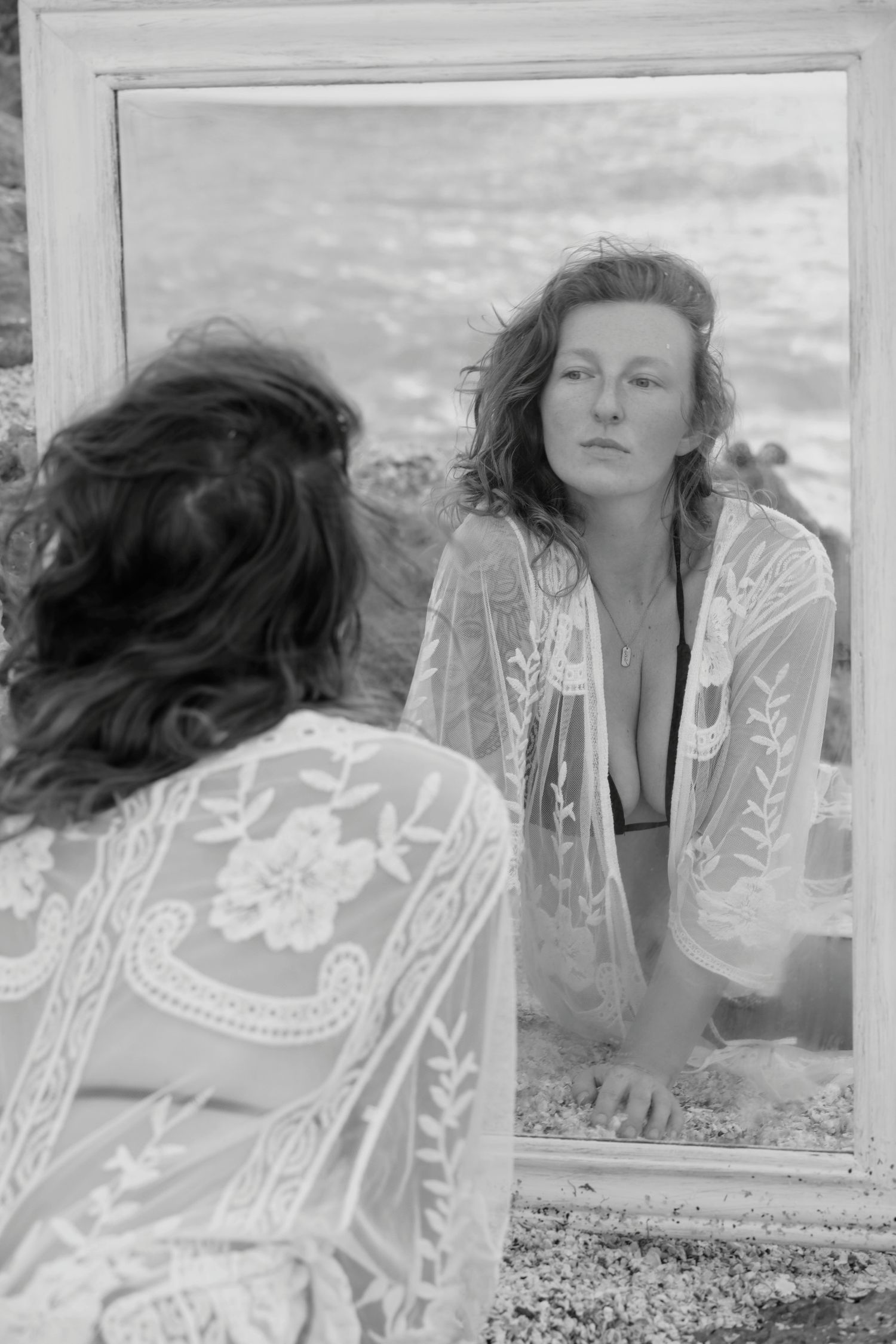Woman in lace cover-up gazes at her reflection in a mirror on a rocky beach in black and white.