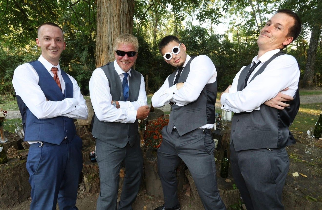 Four groomsmen in gray suits and blue ties pose confidently with crossed arms outdoors at a wedding celebration.
