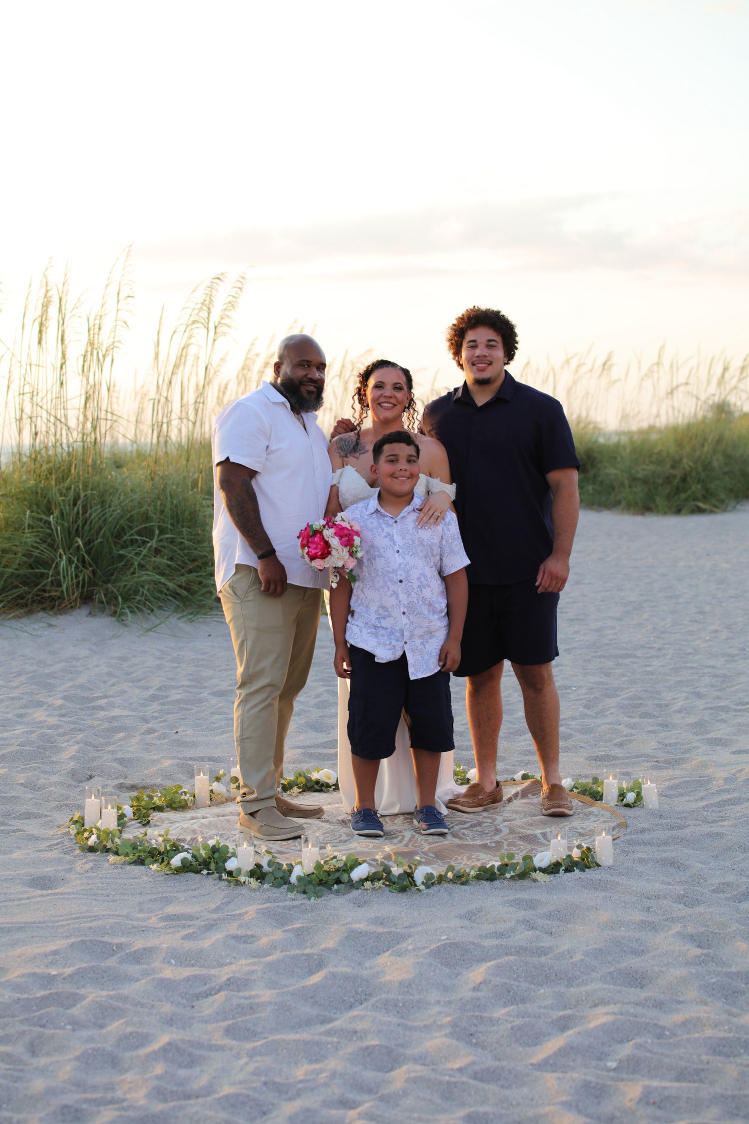 Family of four posing on sandy beach at sunset with floral circle arrangement and sea oats in background.