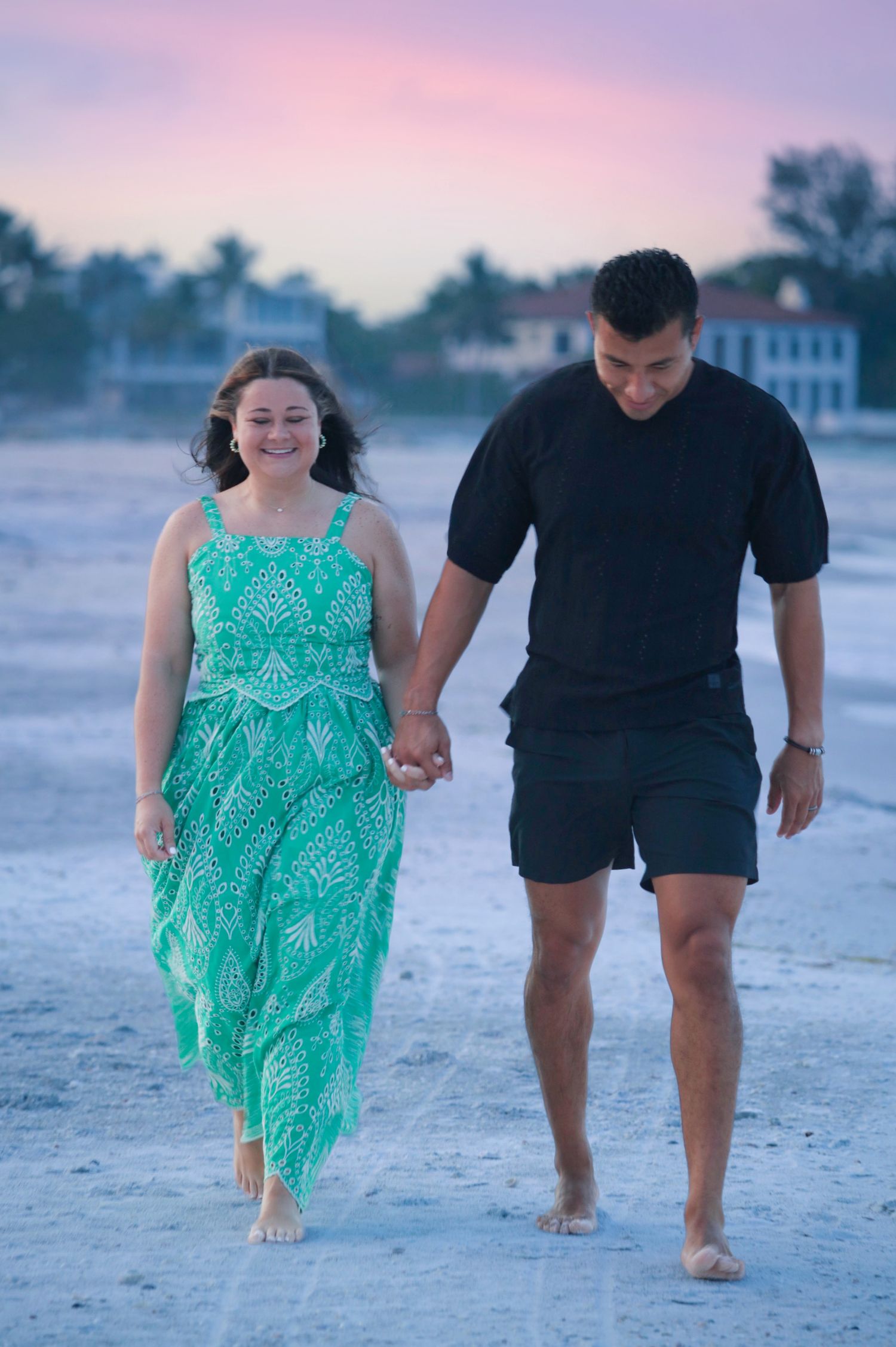 Happy couple holding hands walking barefoot on sandy beach at dusk, woman in green dress.